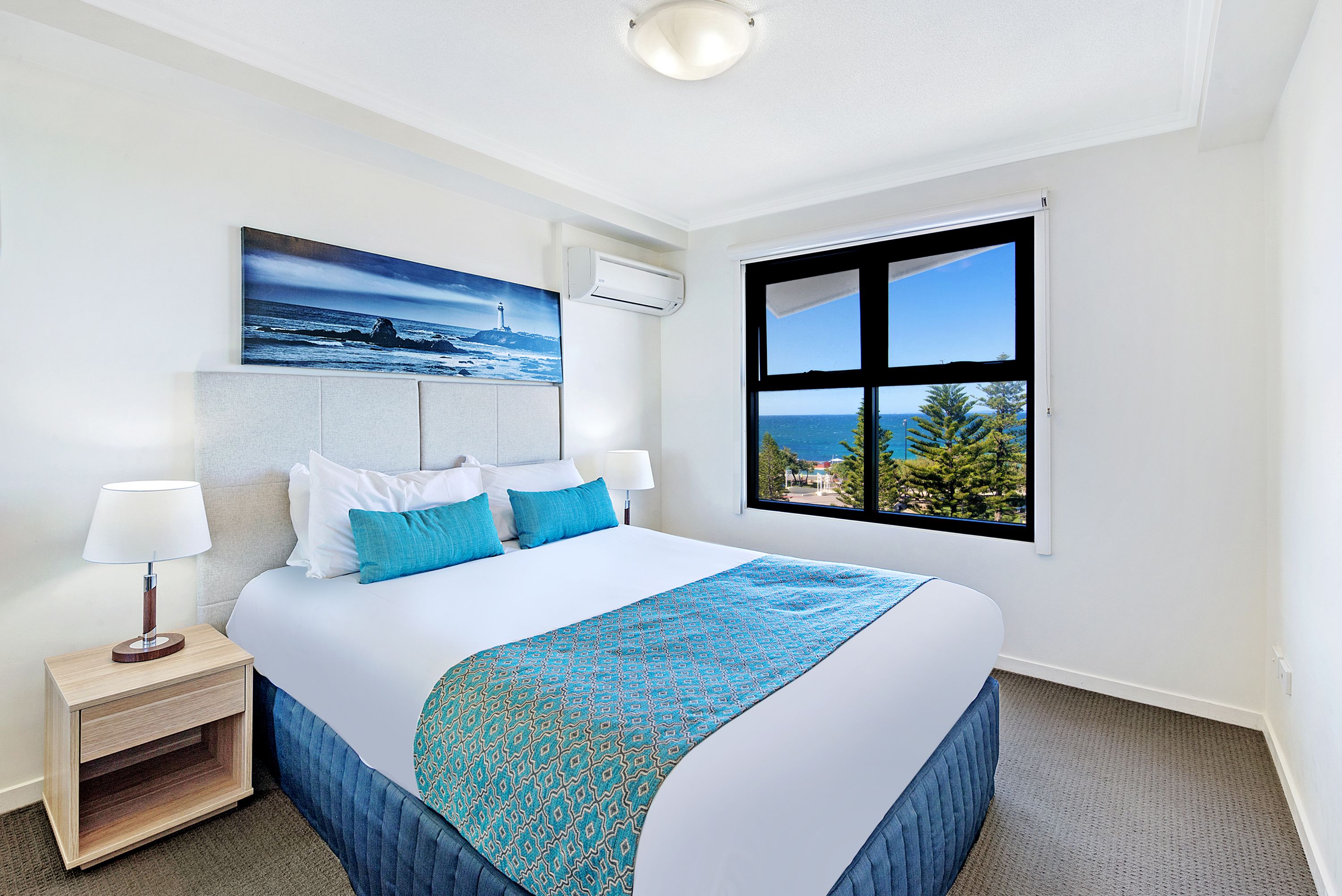 Habitación de hotel con una cama grande, una mesita de noche con una lámpara, un cuadro de un faro y una ventana con vistas a árboles y un edificio.