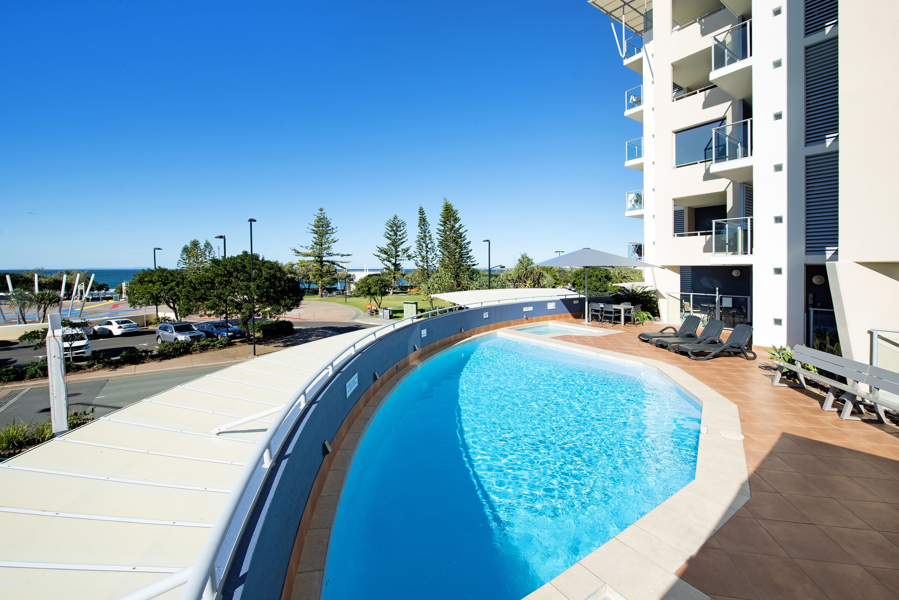 A swimming pool with a curved edge, surrounded by a white building with balconies, and a clear view of the sea in the background.