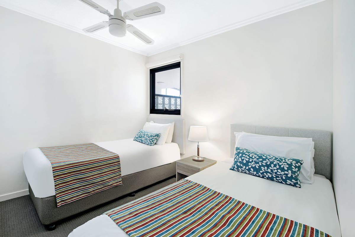 A hotel room with two beds, a ceiling fan, a window with a view, and a nightstand with a lamp.