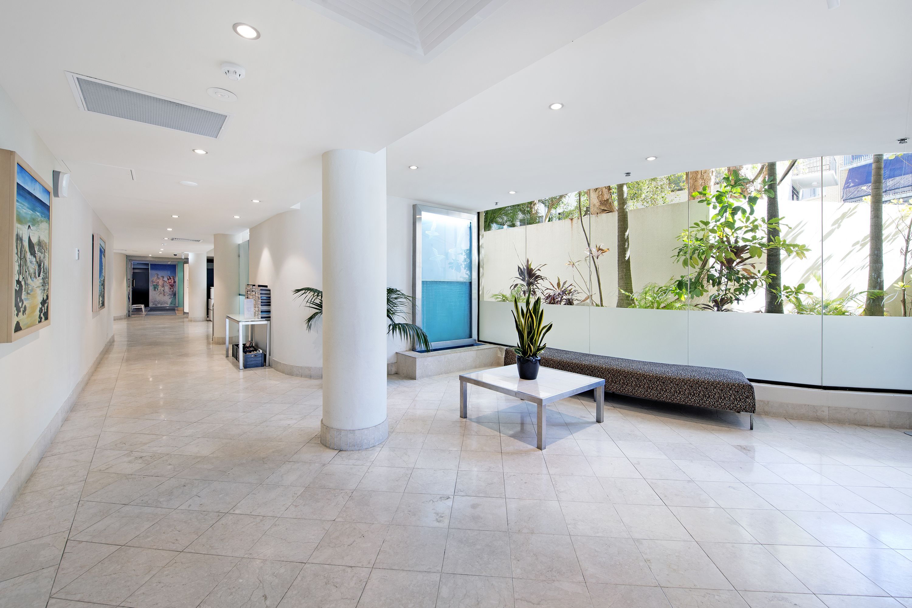 A modern lobby with a tiled floor, a bench, a table with a plant, and a large window with a view of greenery.
