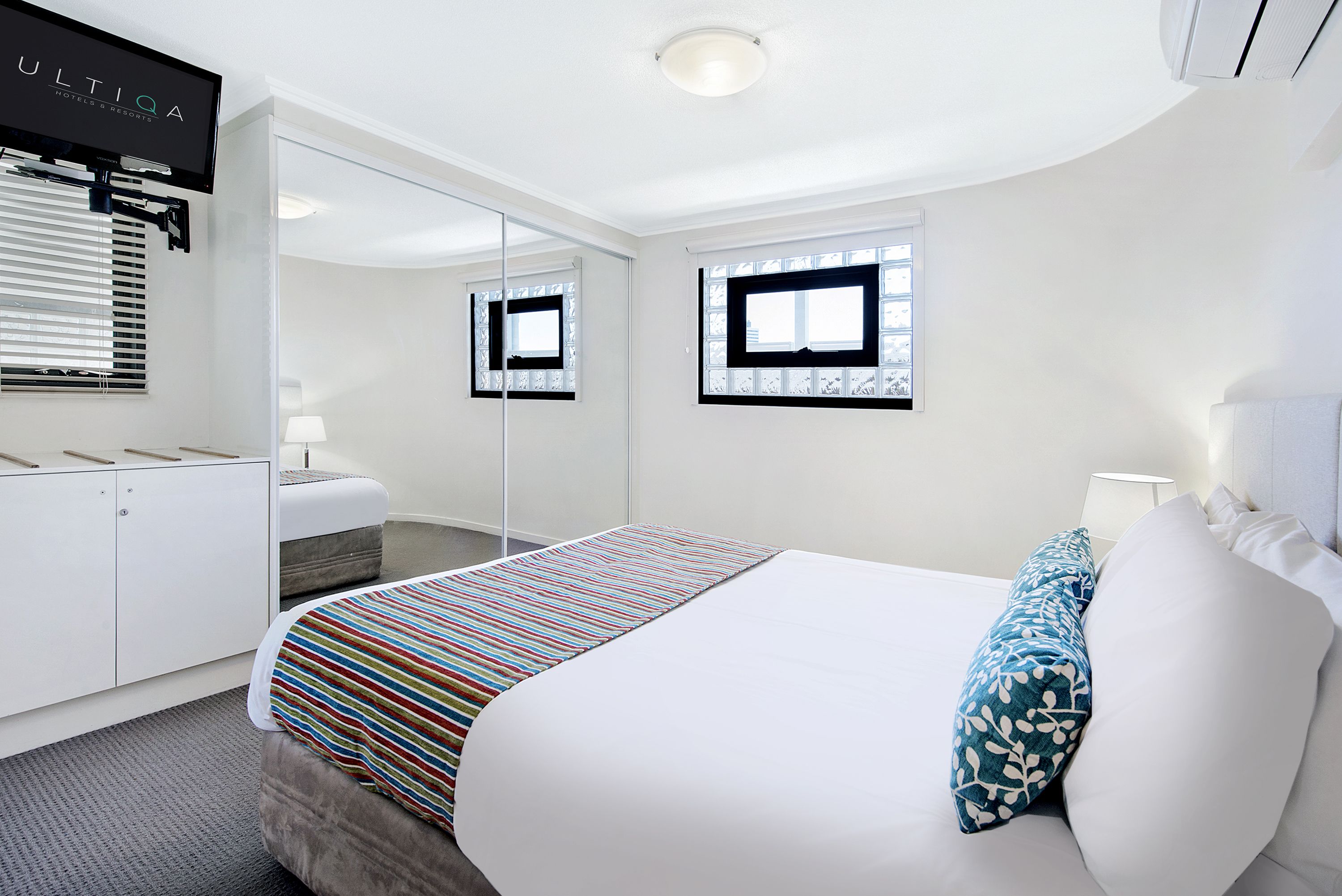 A modern hotel room with a large bed, a bedside table with a lamp, a wardrobe, a mirror, and a TV mounted on the wall.