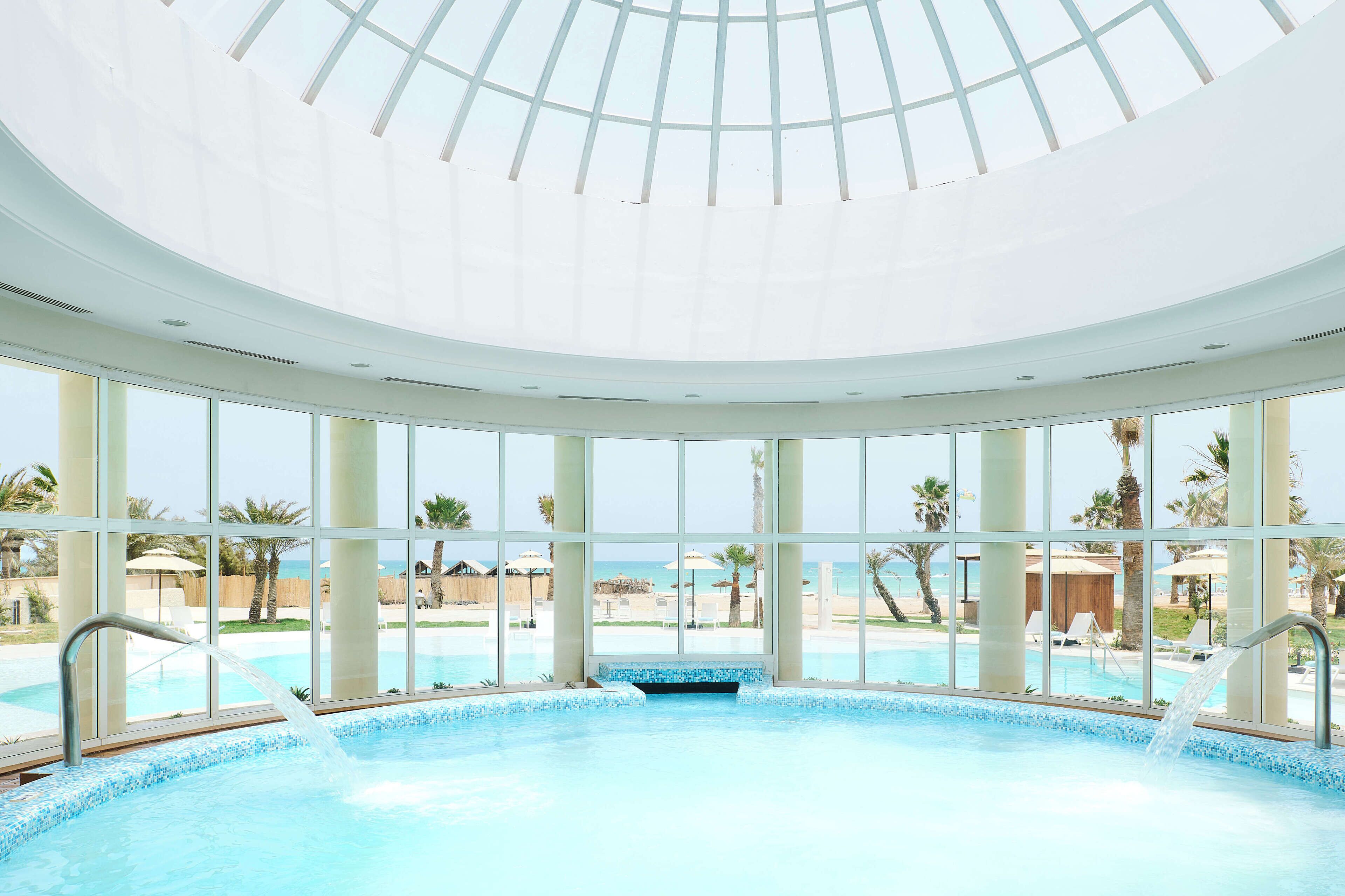 Una amplia piscina interior con vistas despejadas a la playa y palmeras a través de grandes ventanales.