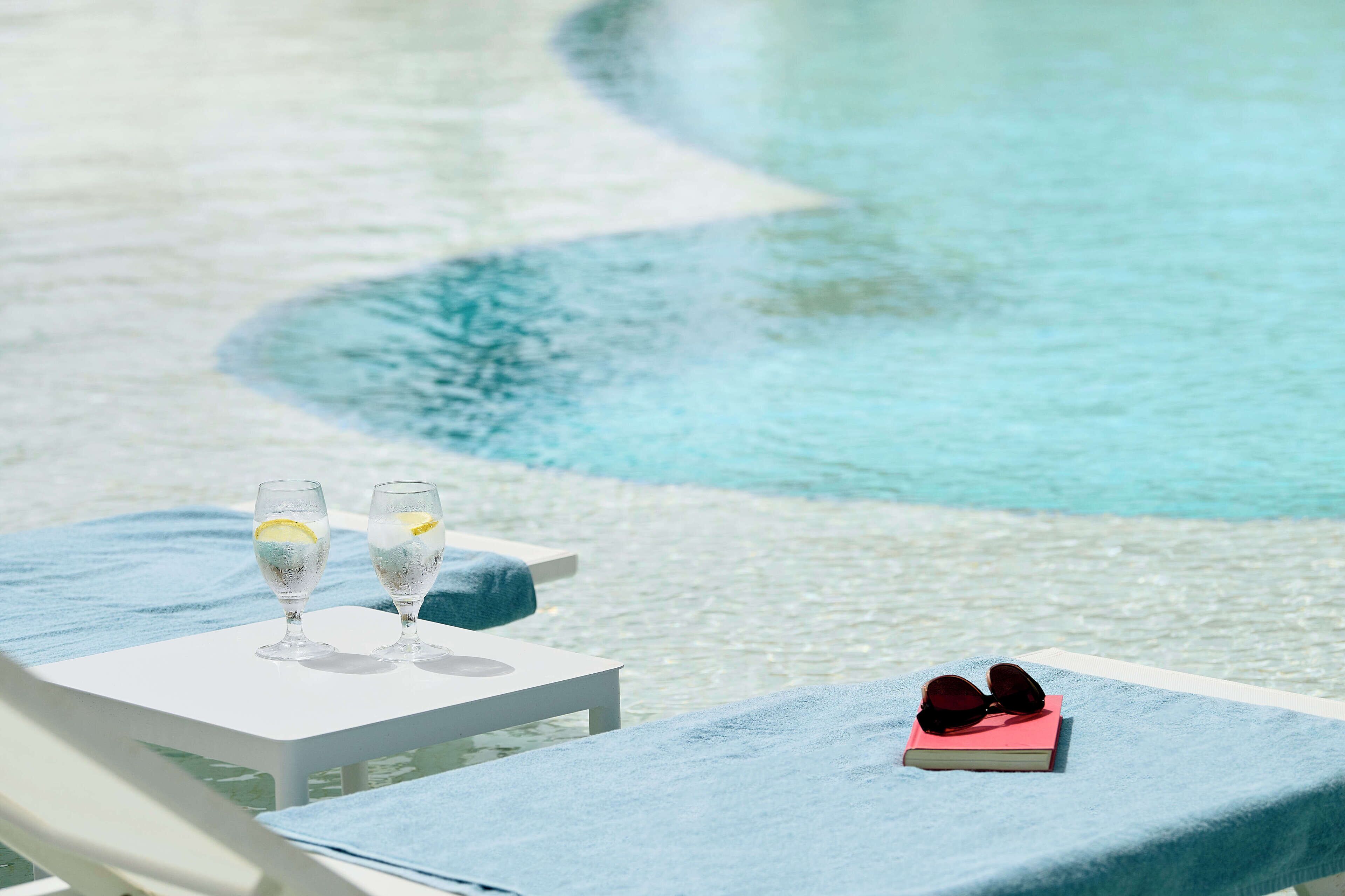 Une scène au bord de la piscine avec deux verres contenant une boisson claire, un livre et une paire de lunettes de soleil posés sur une table.