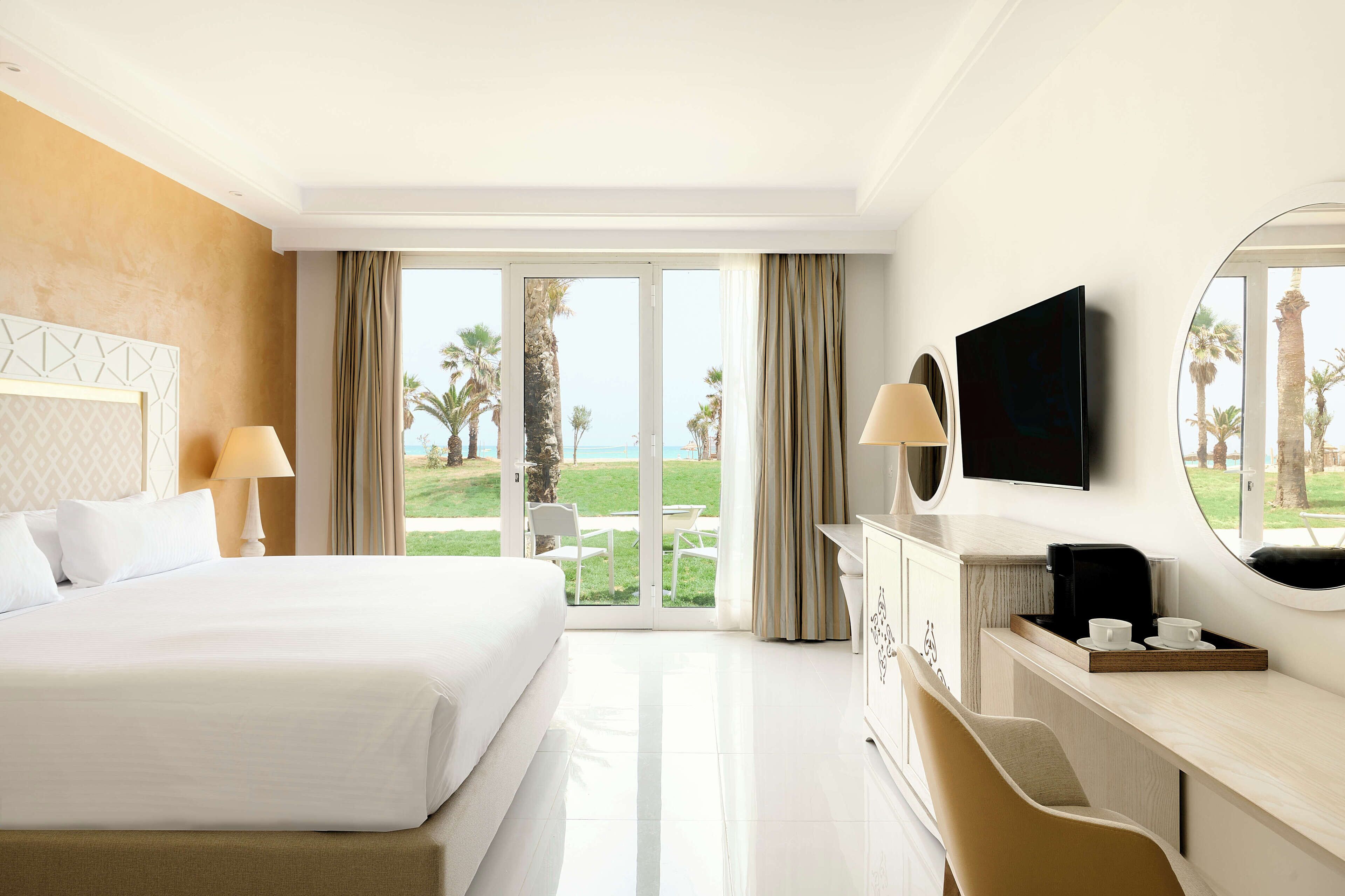A hotel room with a large bed, a desk with a TV, and a view of the beach.