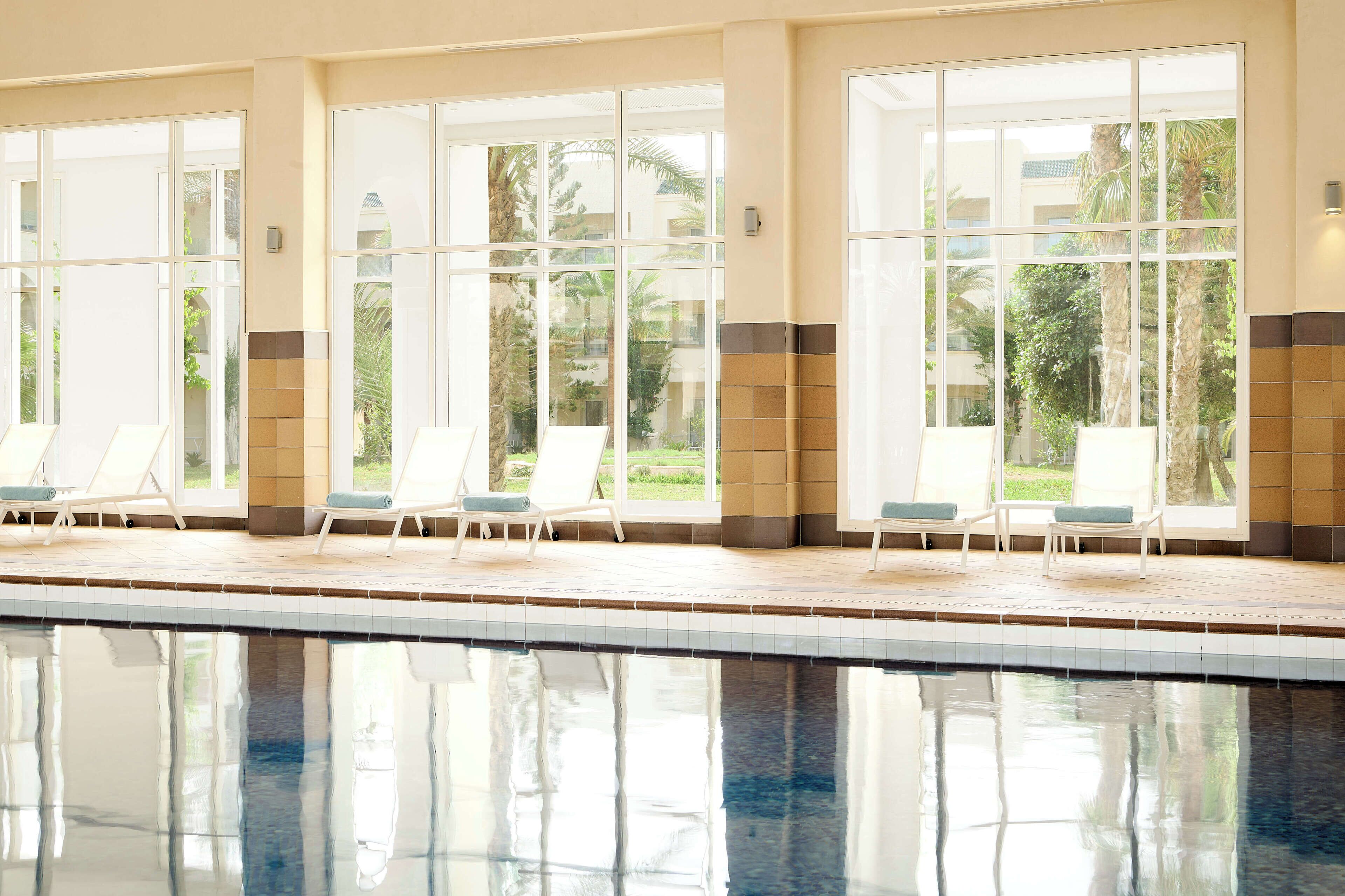 A swimming pool with clear glass windows, lounge chairs, and a view of greenery outside.