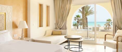 A modern hotel room with a large bed, a sofa, a dining table, and a balcony with a view of the sea.