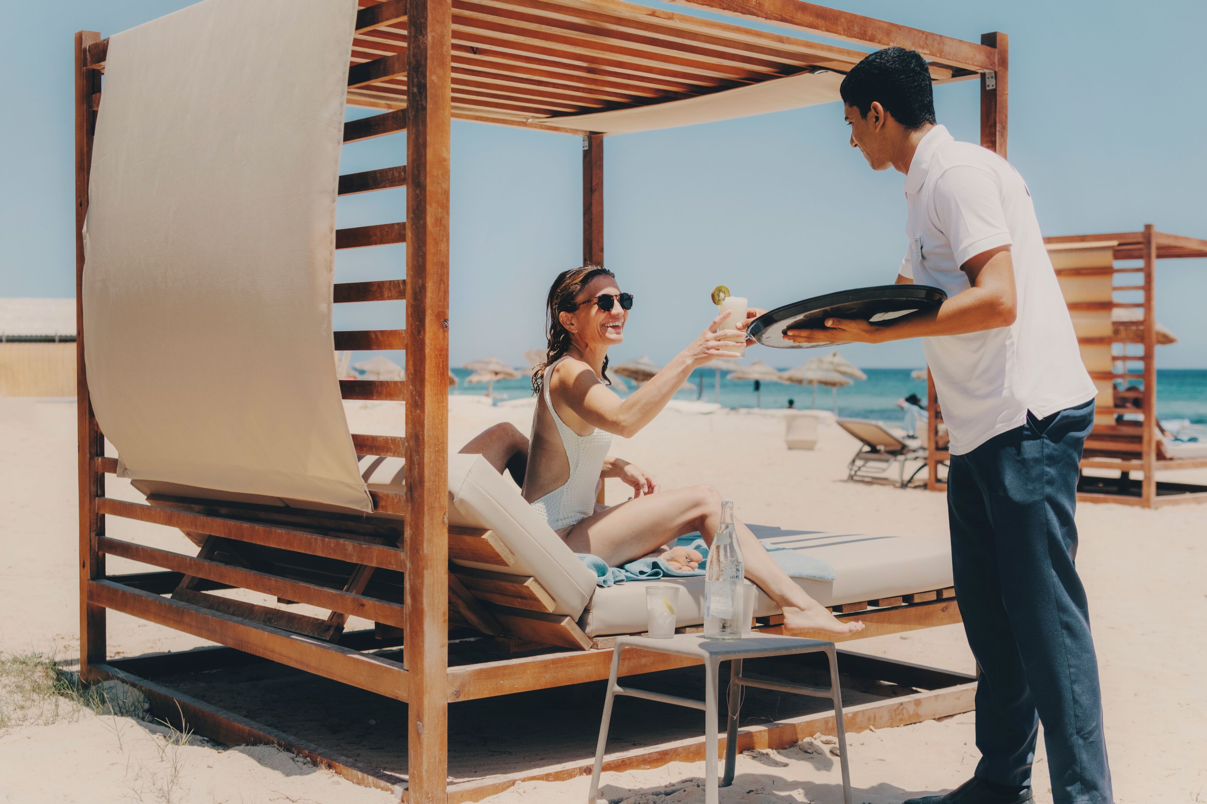 Un cabanon de plage avec une structure en bois et un auvent beige, une chaise longue et une table avec une bouteille et un verre.