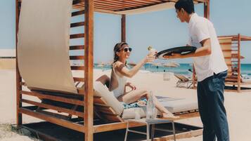 Un cabanon de plage avec une structure en bois et un auvent beige, une chaise longue et une table avec une bouteille et un verre.