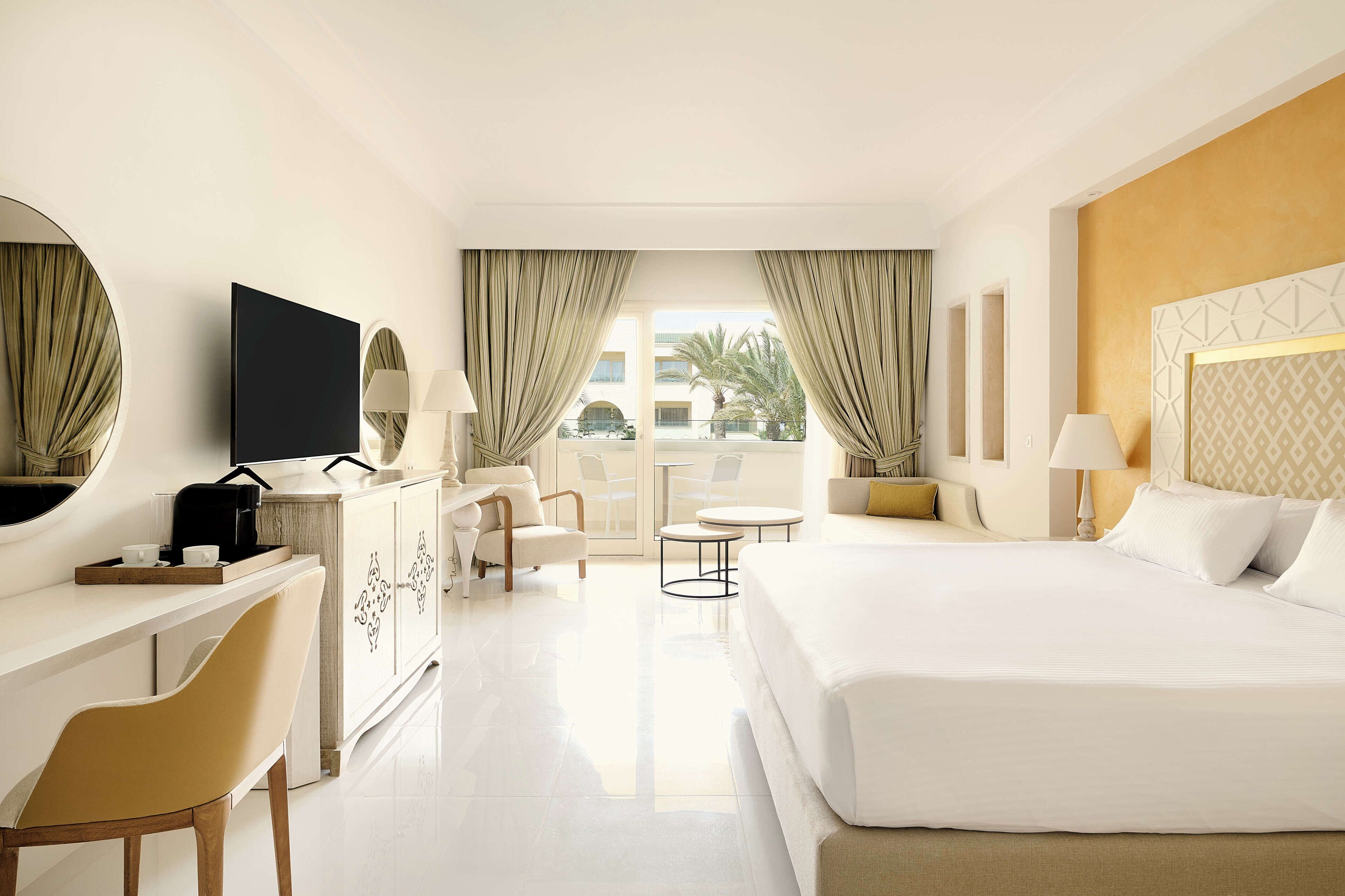Quarto de hotel com uma cama grande, uma escrivaninha com televisão, uma cadeira, uma mesinha e vista para palmeiras pela janela.