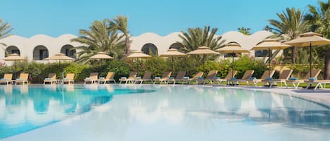 A pool area with lounge chairs and umbrellas, surrounded by palm trees and a building with arched windows.