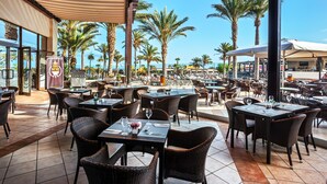 An outdoor restaurant with wicker chairs and tables, palm trees, and a view of the beach. - Occidental Jandía Mar - All Inclusive (Pajara)
