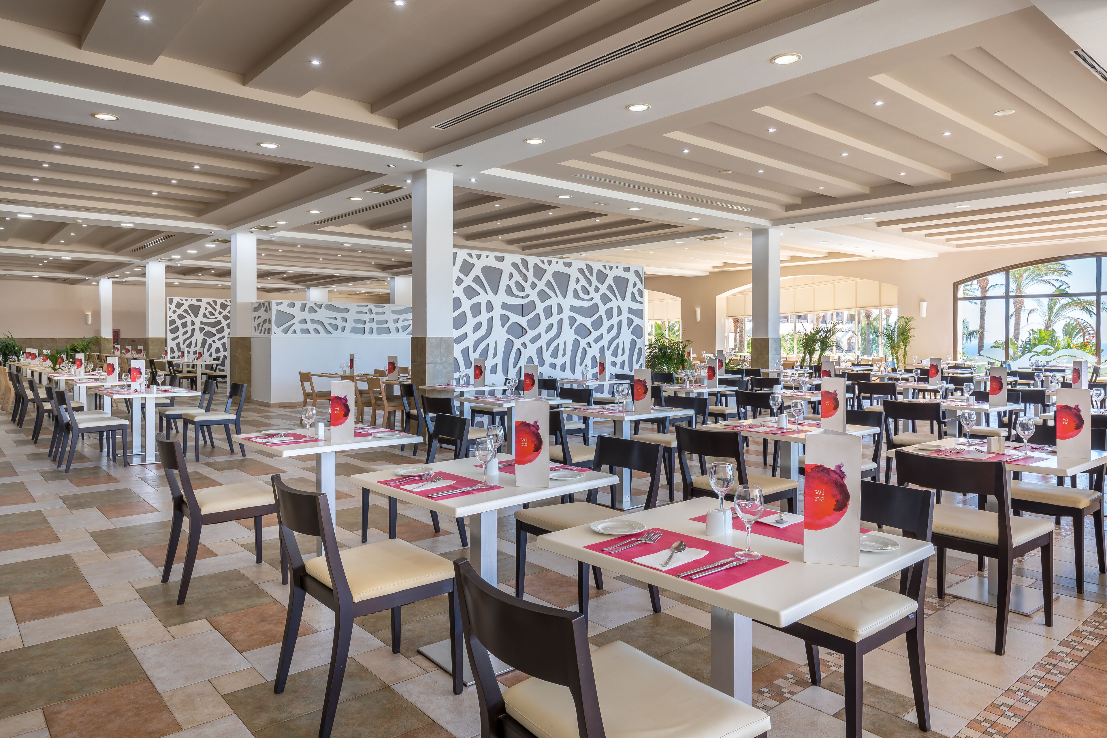 A spacious dining area with multiple tables and chairs, a modern ceiling design, and large windows allowing natural light.