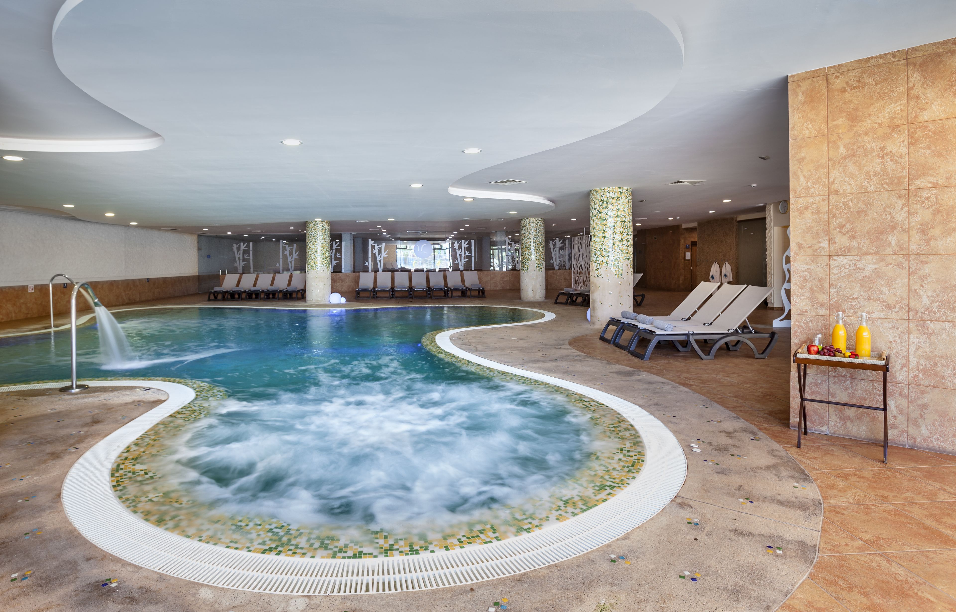 A large indoor swimming pool with a hot tub in the center, surrounded by lounge chairs and a small table with bottles.