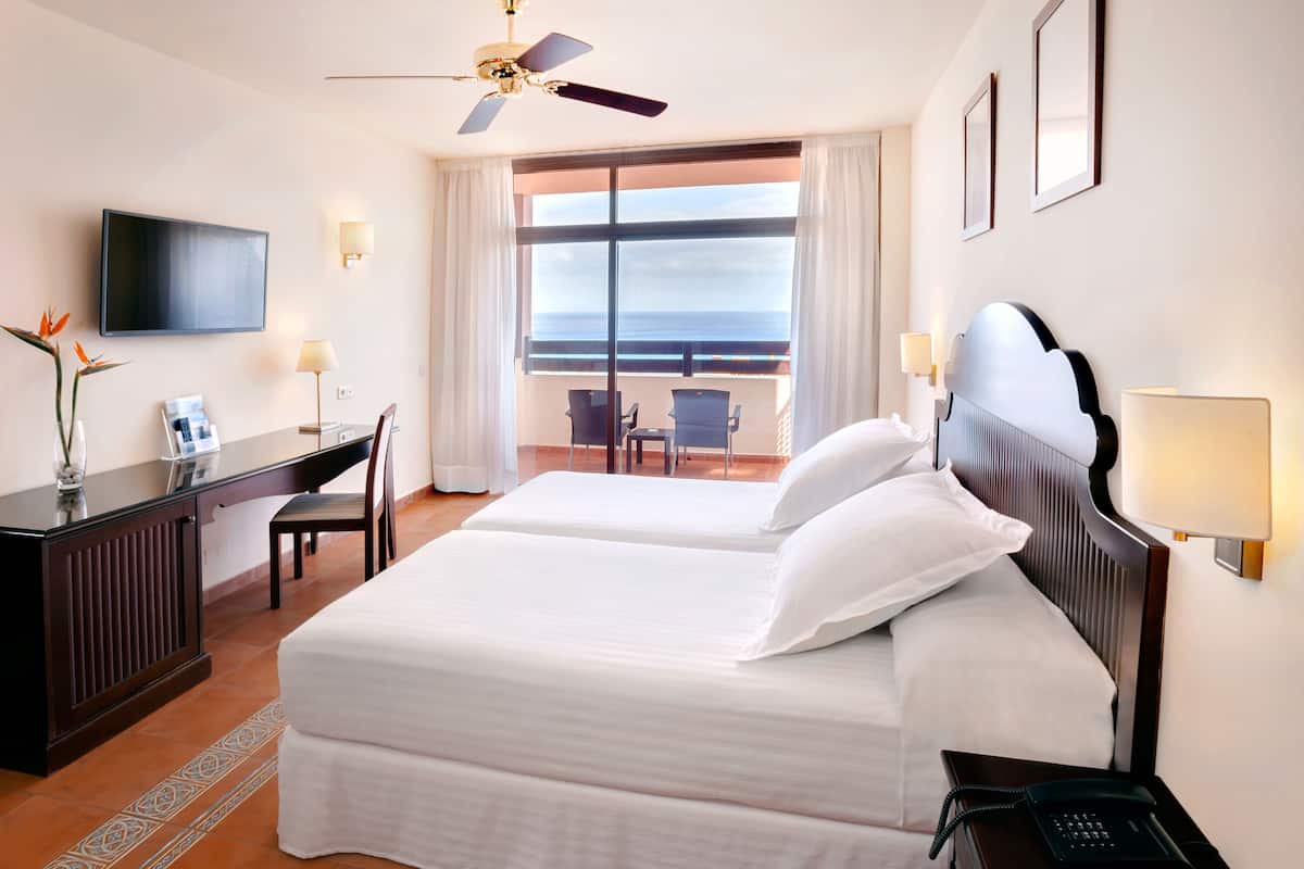 Ein Hotelzimmer mit zwei Betten, einem Schreibtisch, einem Stuhl, einem Fernseher und einem Balkon mit Meerblick.