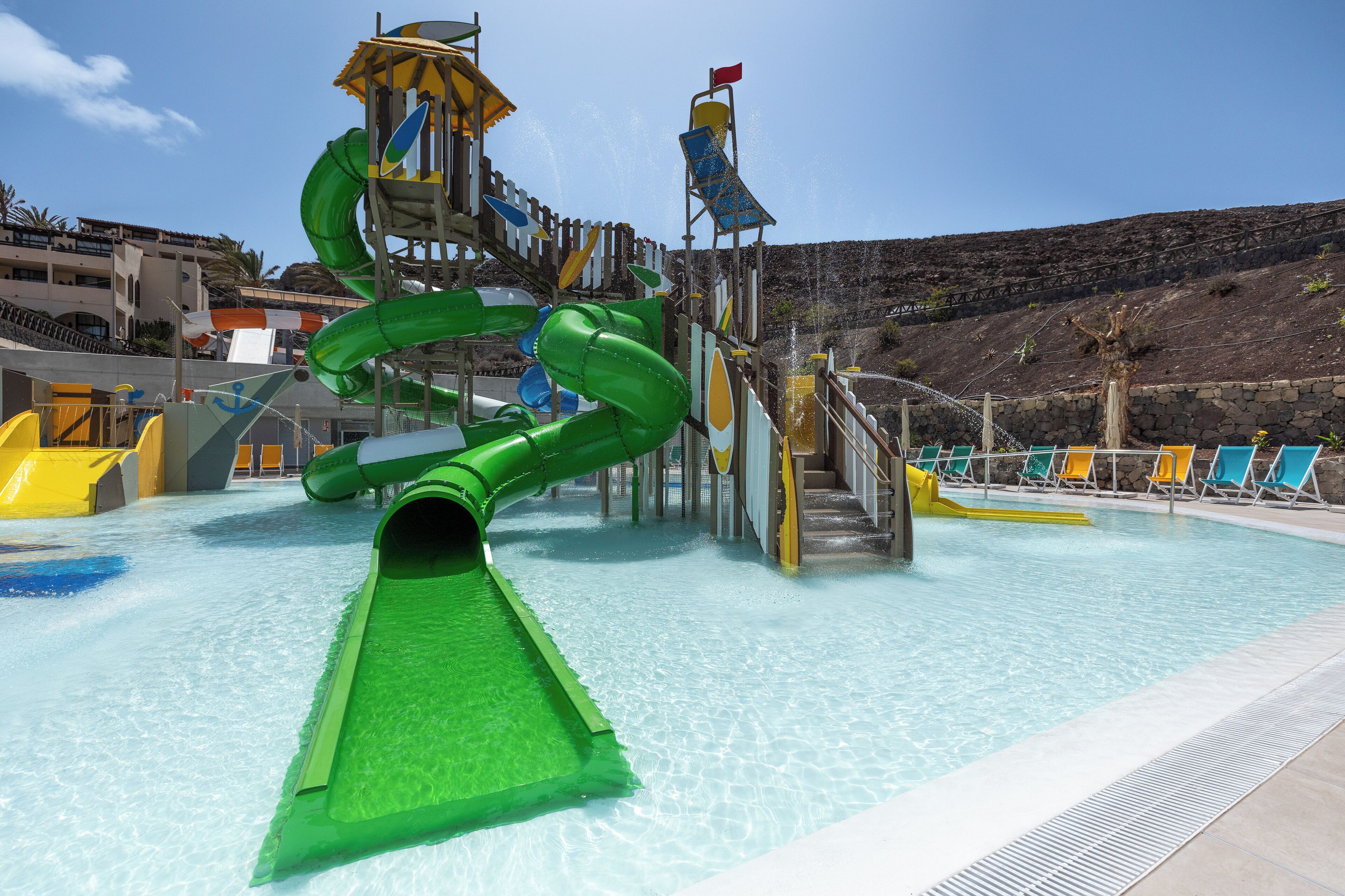 Parque acuático con un tobogán verde, toboganes amarillos y azules, y una zona de piscina con sillones de descanso.