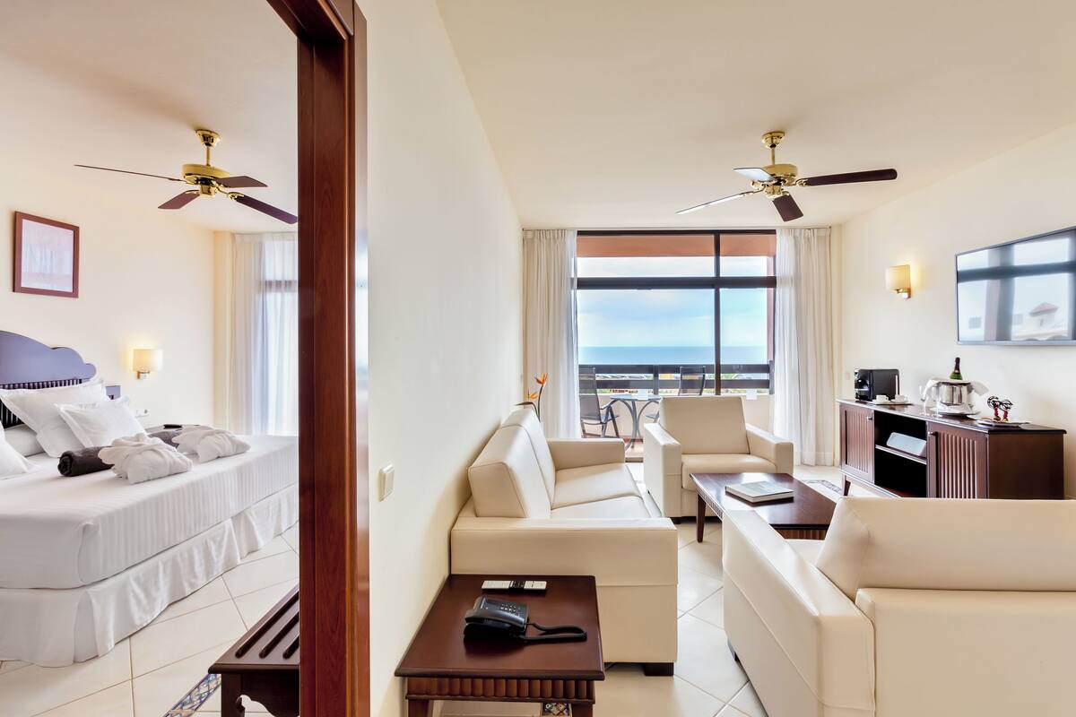 Ein Hotelzimmer mit einem Bett, einem Sofa, einem kleinen Tisch und einem Schreibtisch mit einem Computer. Es gibt Deckenventilatoren, ein Fenster mit Blick und einen Fernseher an der Wand.