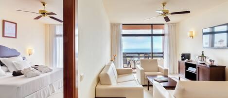 A hotel room with a bed, a sofa, a small table, and a desk with a computer. There are ceiling fans, a window with a view, and a TV on the wall.