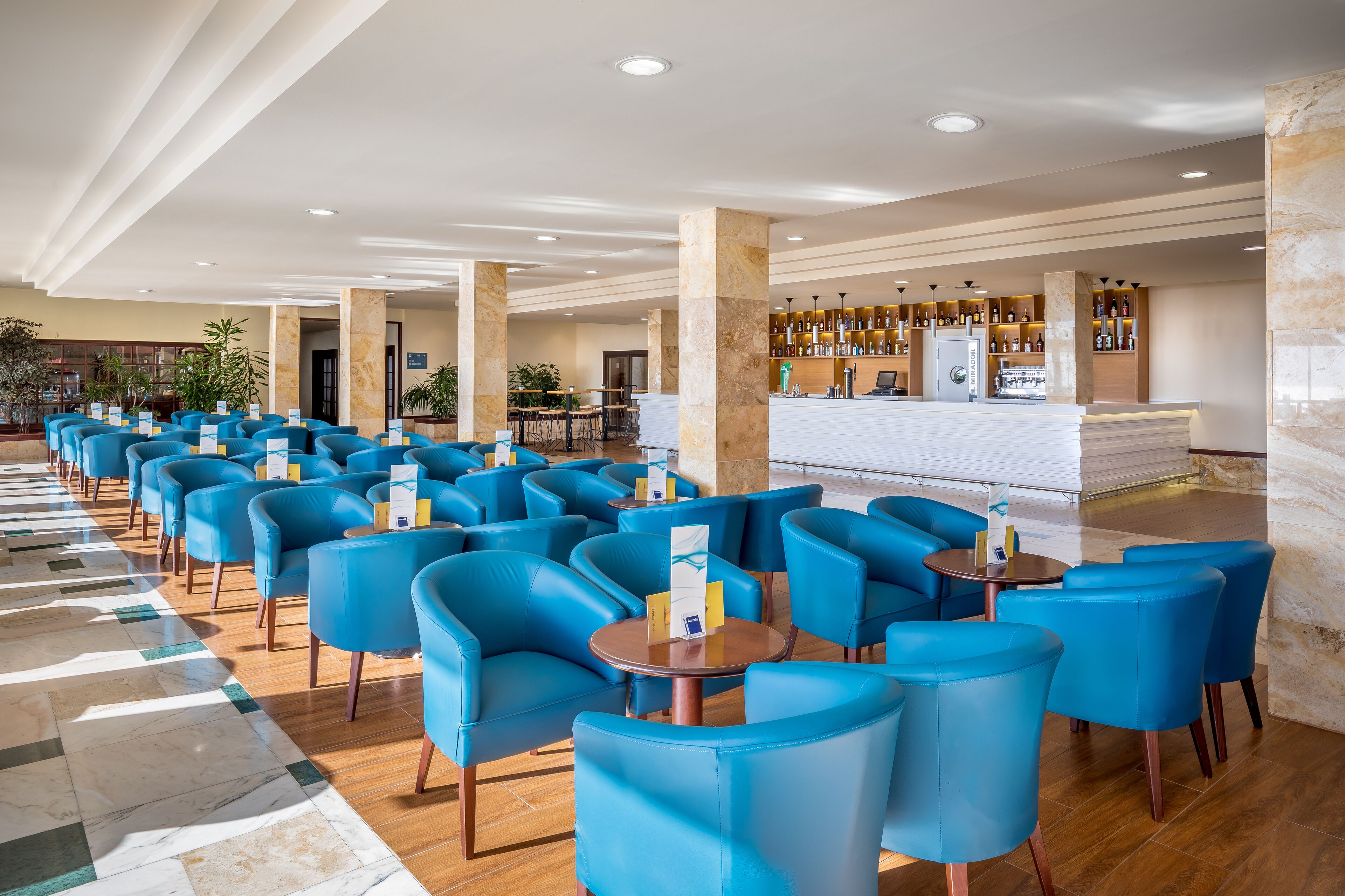 A hotel lobby with rows of blue chairs, a bar area with bottles, and a reception desk.