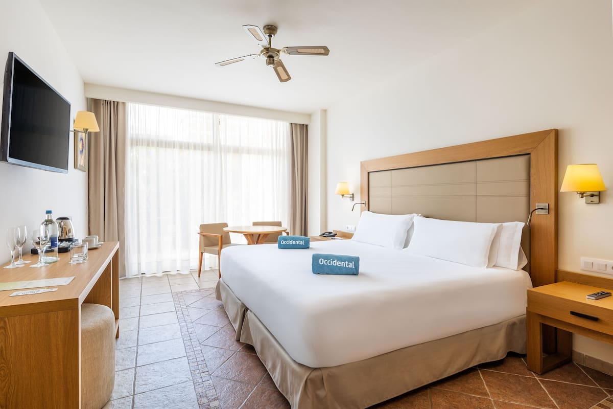 Ein Hotelzimmer mit Bett, Schreibtisch, Sessel, Fernseher und Deckenventilator.