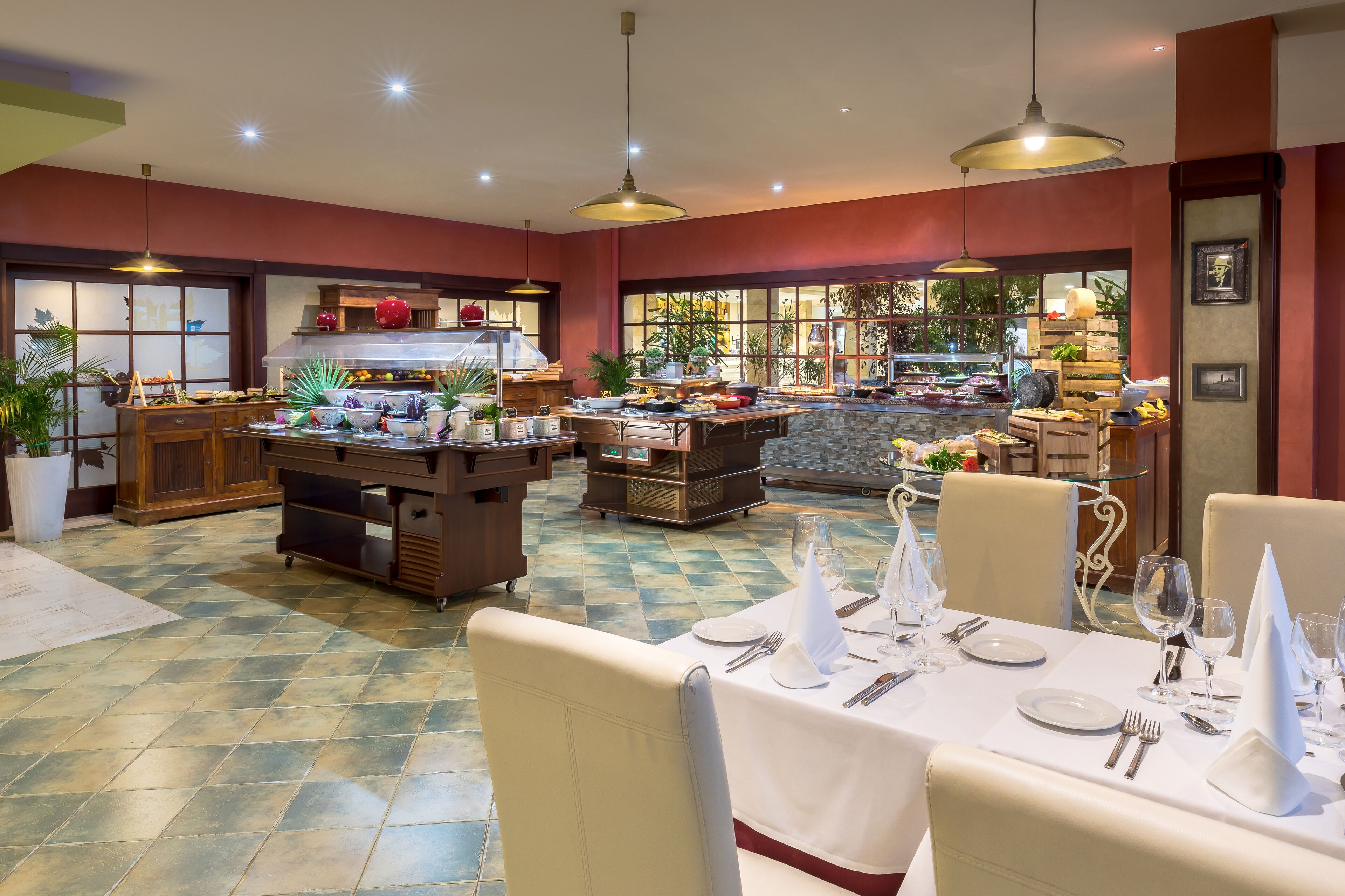 A spacious dining area with multiple tables set for service, a kitchen in the background, and a view of the outdoors through large windows.