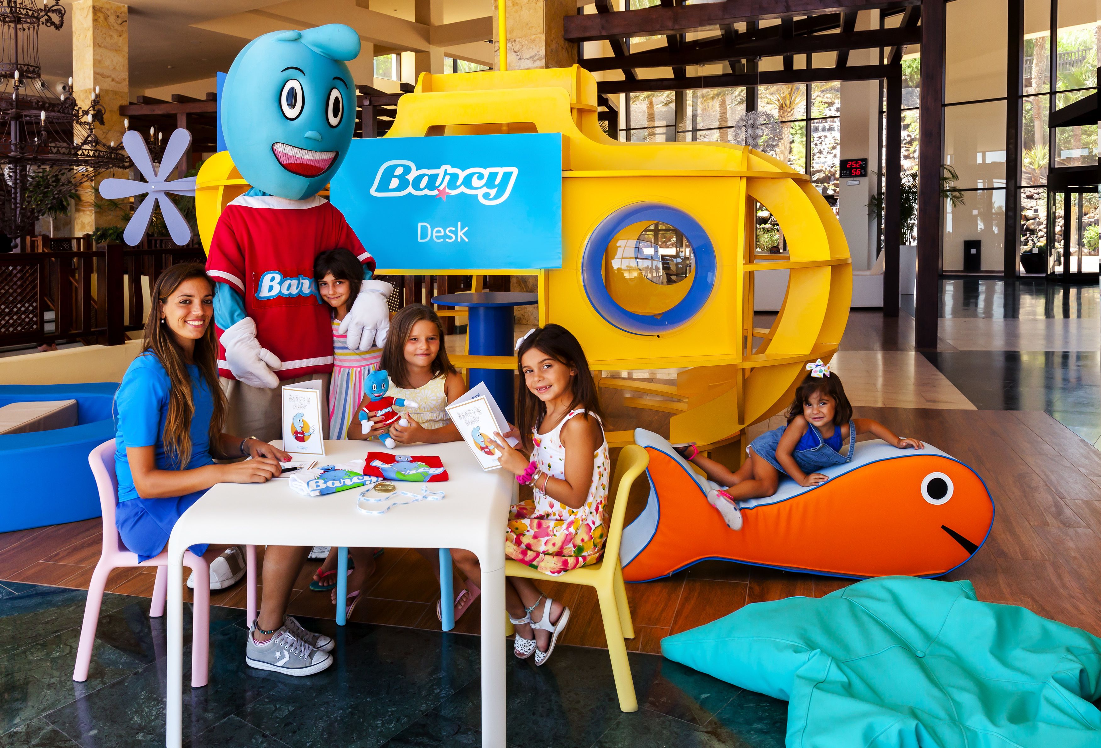 A group of children and an adult at a table with a mascot, surrounded by colorful play structures and a 'Barcy Desk' sign.