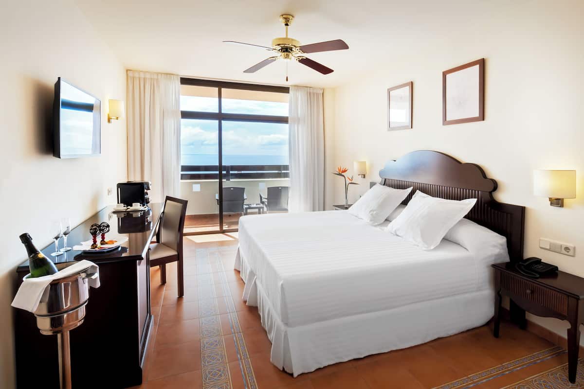 Una habitación de hotel con cama, escritorio con una botella de champán, una silla, televisión y balcón con vistas.