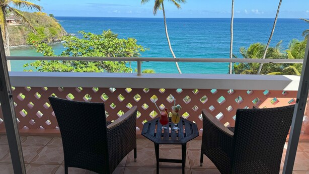 A balcony with wicker chairs and a small table, overlooking a coastal view with palm trees and the ocean. - Blue Haven Hotel (Scarborough)