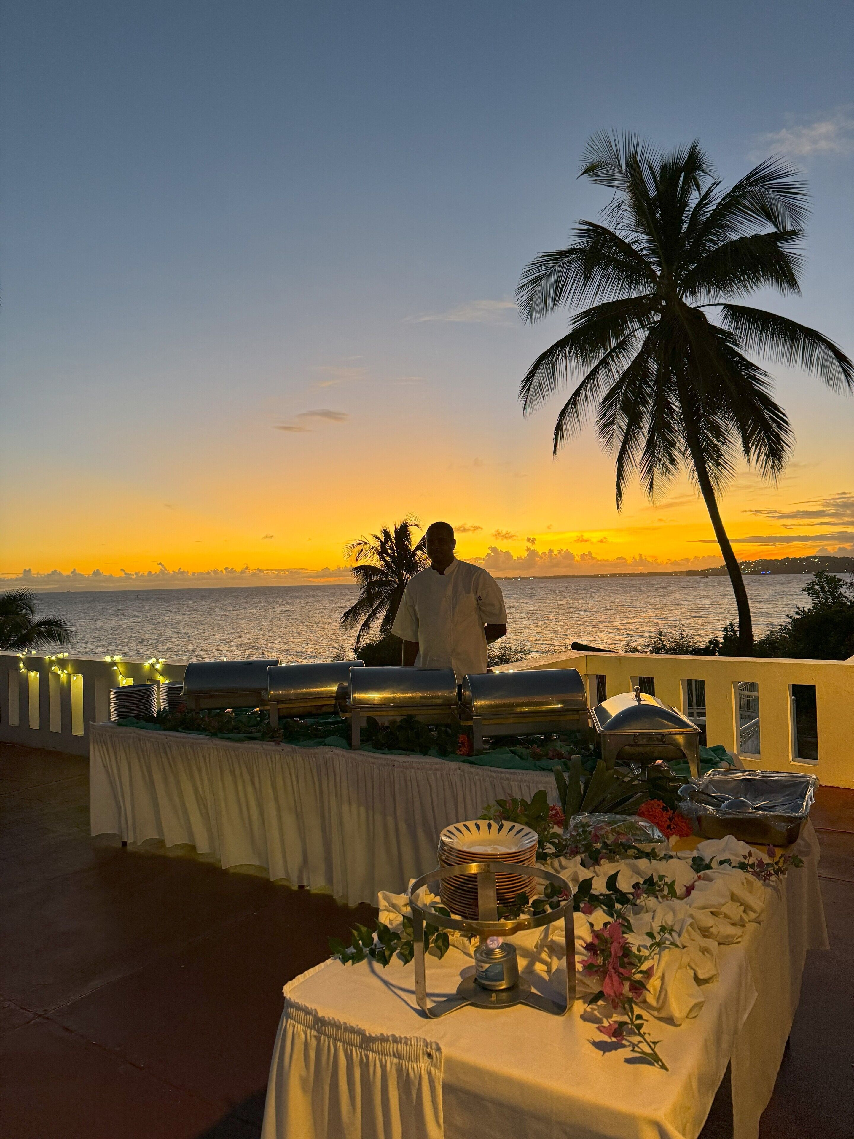 A buffet table set up on a terrace overlooking a sunset view, with a palm tree and a person in the background.