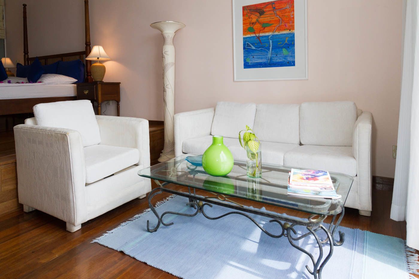 A living room with a white sofa, a white armchair, a glass coffee table with a green vase and a book, and a tall white lamp.
