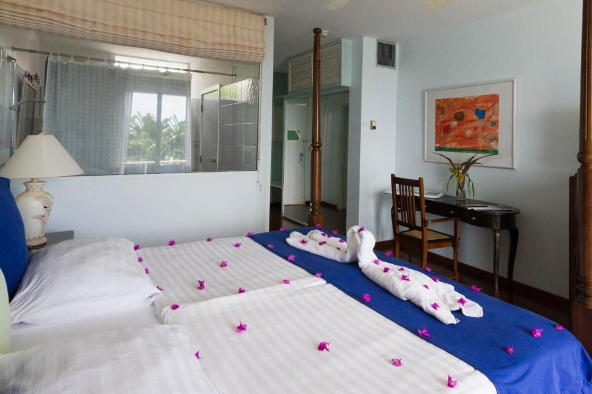 A hotel room with two beds, a desk, a chair, a lamp, a window with curtains, and a painting on the wall.