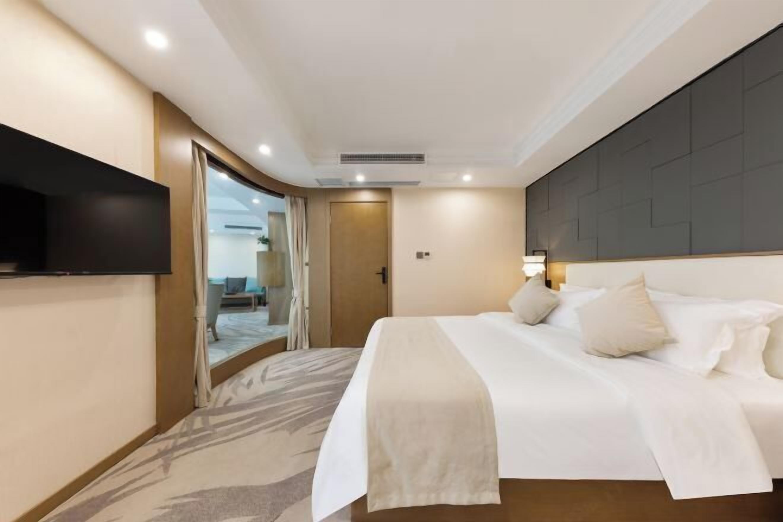 A hotel room with a bed, a flat-screen TV mounted on the wall, a door leading to a bathroom, and a view of the outside.