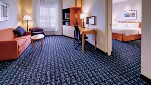 A hotel room with a blue patterned carpet, a red sofa, a desk with a lamp, a chair, a bed, and a painting on the wall. - Fairfield Inn & Suites by Marriott Wausau (Schofield)