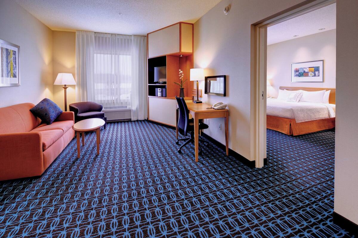 Une chambre d’hôtel avec un tapis à motifs bleus, un canapé rouge, un bureau avec une lampe, une chaise, un lit et un tableau accroché au mur.
