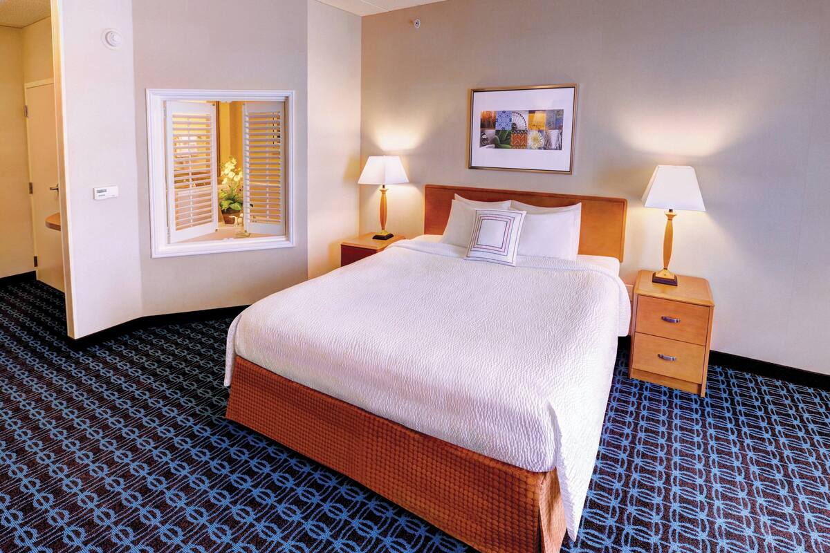 Une chambre d’hôtel avec un grand lit, deux tables de chevet avec des lampes, une fenêtre avec des stores, un tableau encadré au mur et une moquette à motifs.