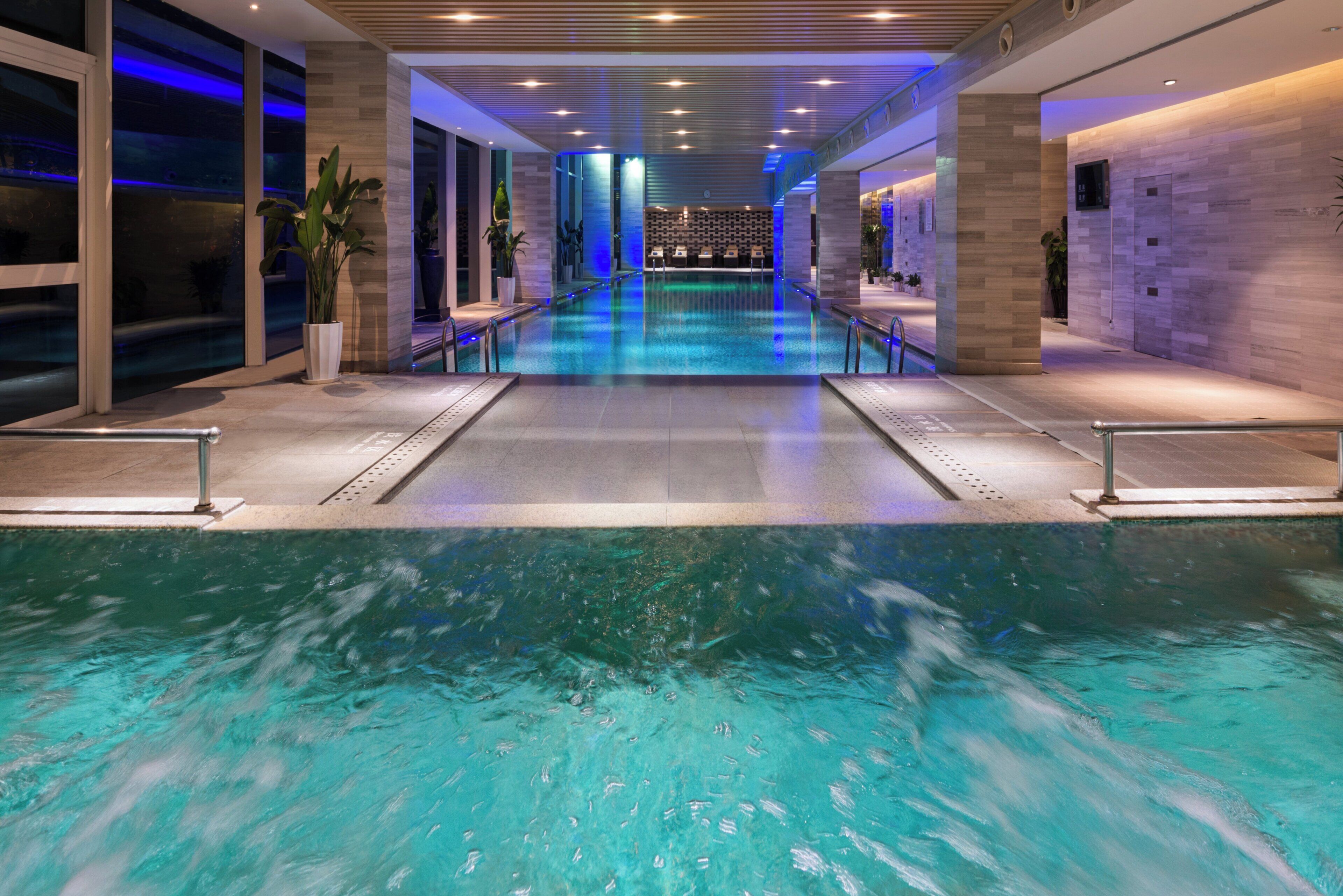 A modern indoor swimming pool with clear water, surrounded by a tiled walkway and potted plants.