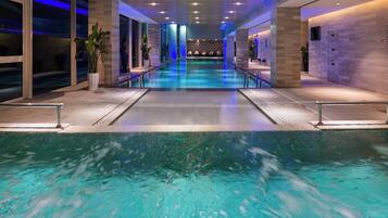 A modern indoor swimming pool with clear water, surrounded by a tiled walkway and potted plants.