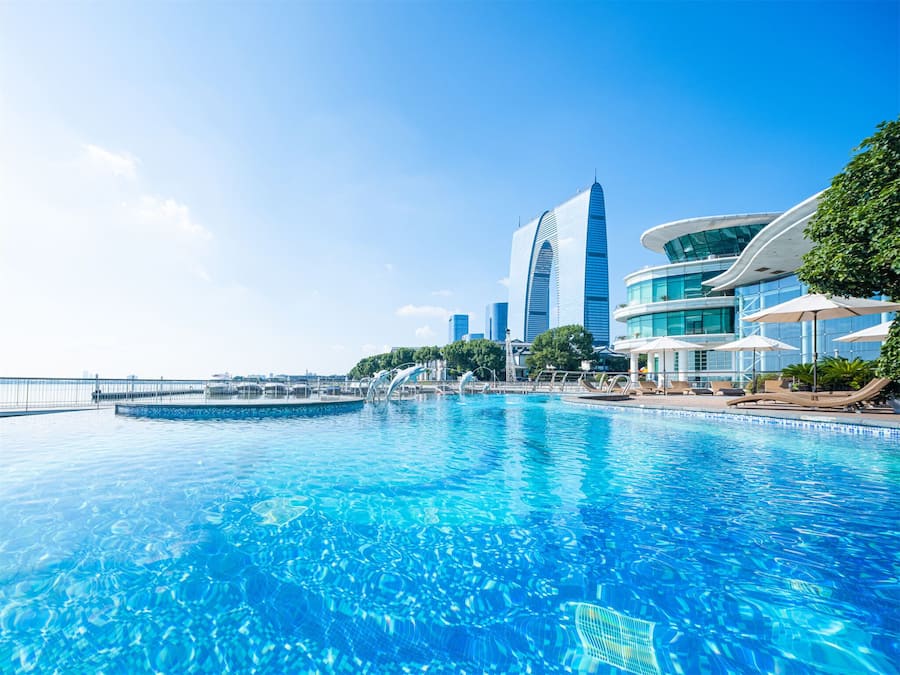A swimming pool with clear blue water, surrounded by modern buildings and a view of the sea.