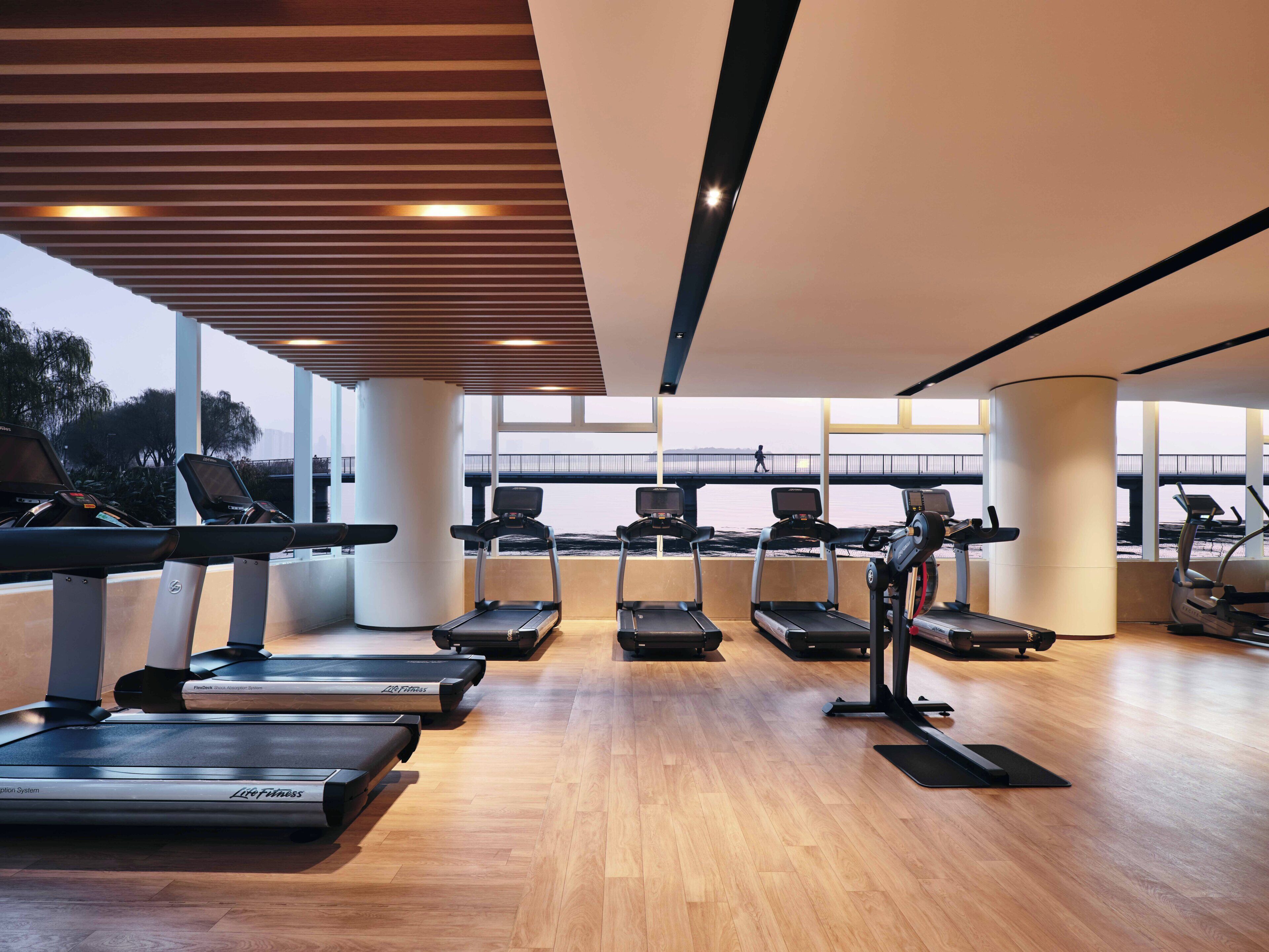 Un gimnasio con cintas de correr, una bicicleta estática y una máquina de remo, con amplias ventanas que ofrecen vistas a un puente y árboles.