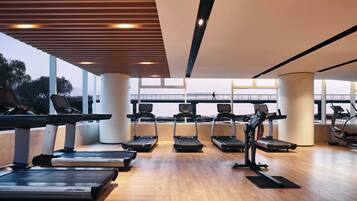 Un gimnasio con cintas de correr, una bicicleta estática y una máquina de remo, con amplias ventanas que ofrecen vistas a un puente y árboles.
