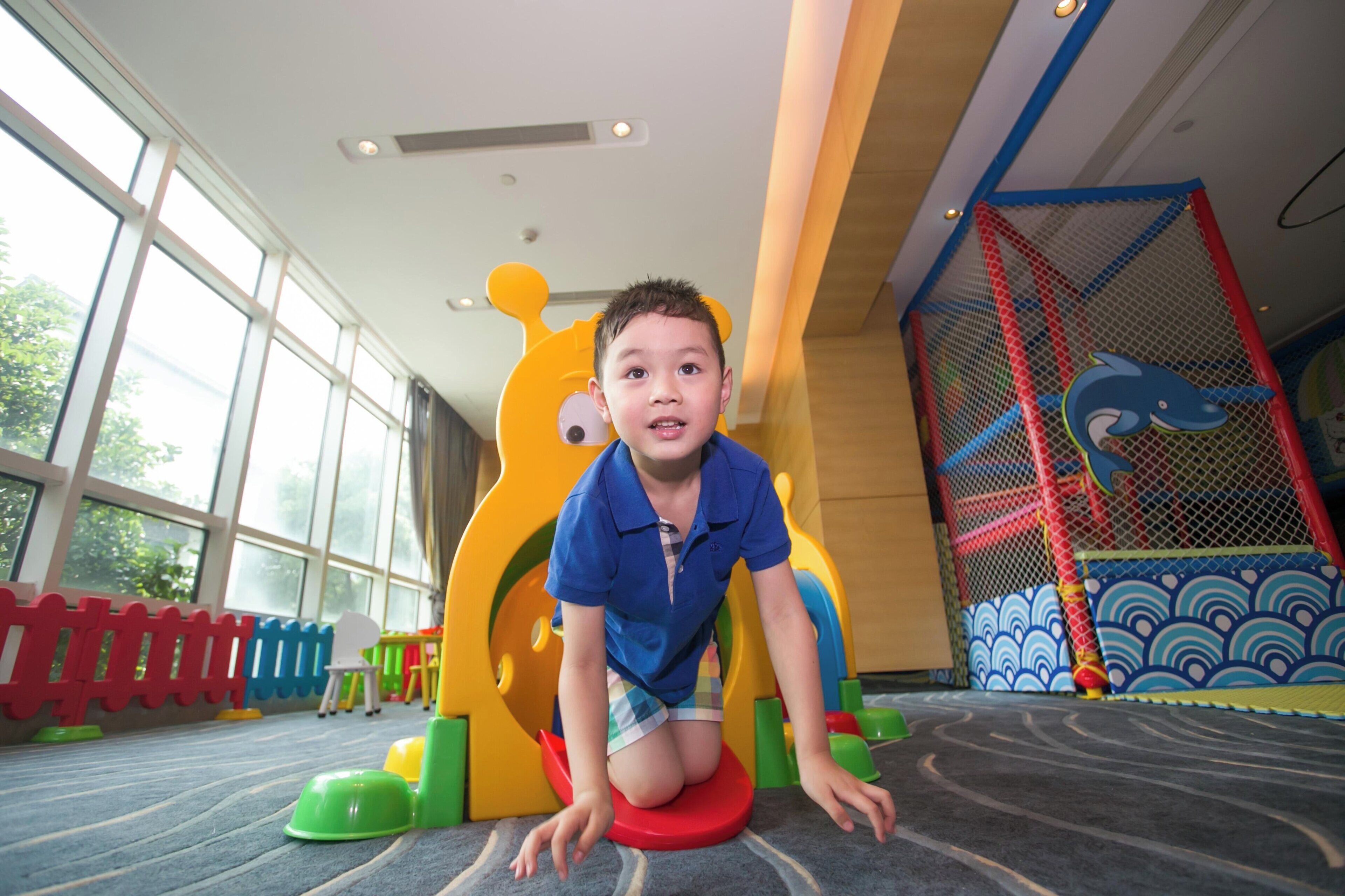Niño jugando en un suelo acolchado con estructuras de juego coloridas, incluyendo un tobogán y una zona de escalada.