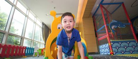 Niño jugando en un suelo acolchado con estructuras de juego coloridas, incluyendo un tobogán y una zona de escalada.