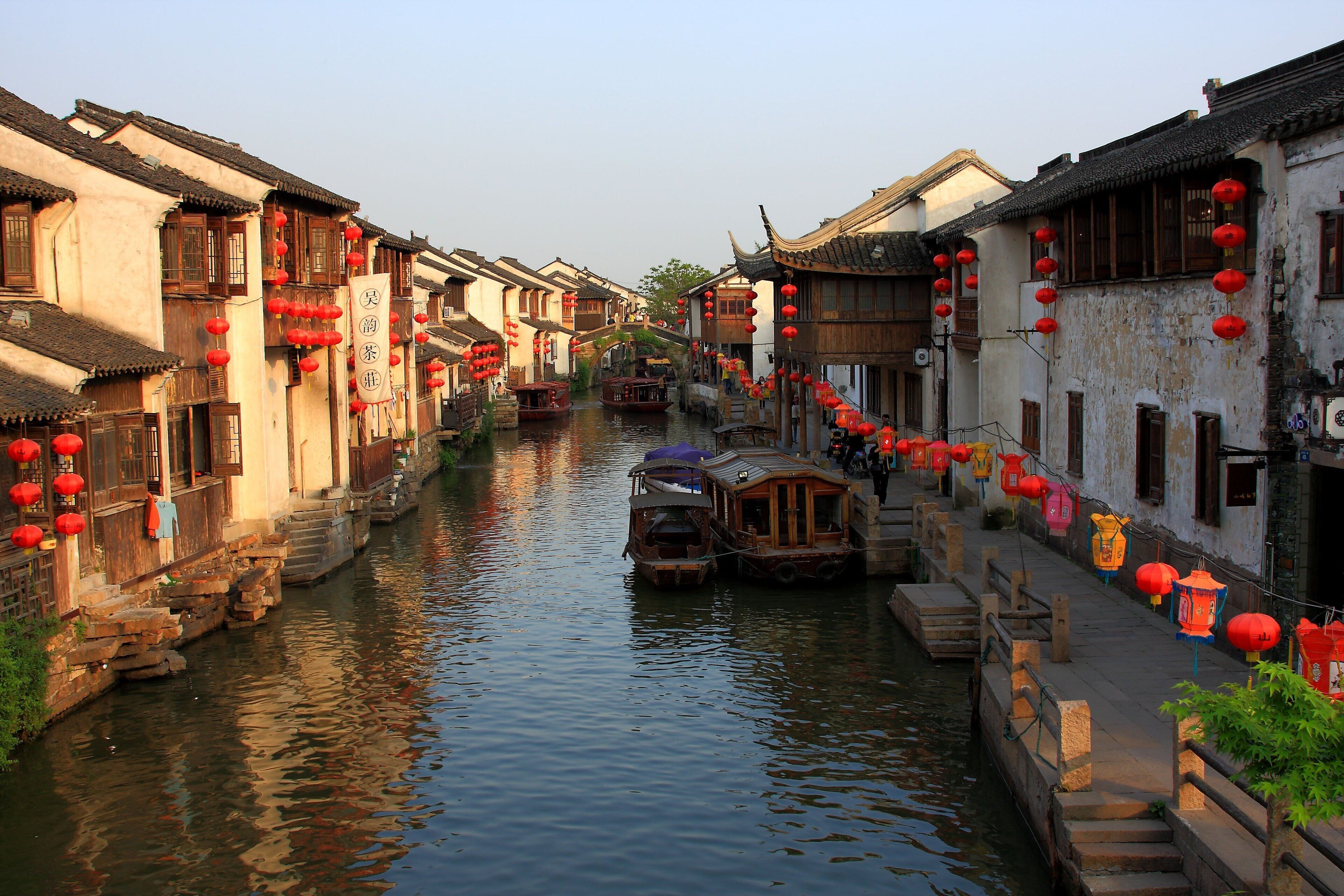 Un étroit canal bordé de bâtiments traditionnels ornés de lanternes rouges, de bateaux en bois et d’escaliers en pierre menant à l’eau.