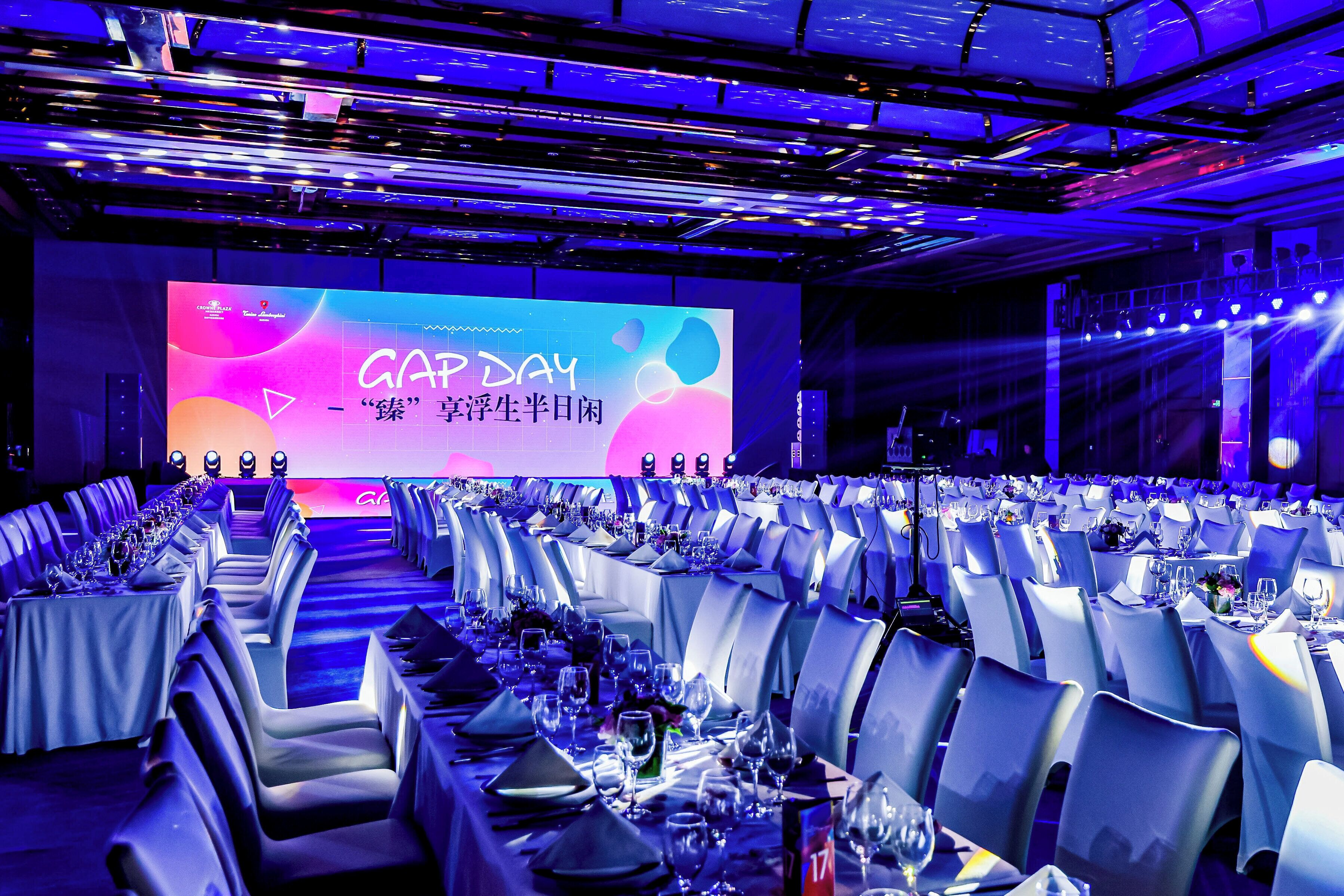 Un salón de conferencias con filas de mesas dispuestas para un evento formal, una pantalla grande mostrando texto y un escenario con equipo de iluminación.