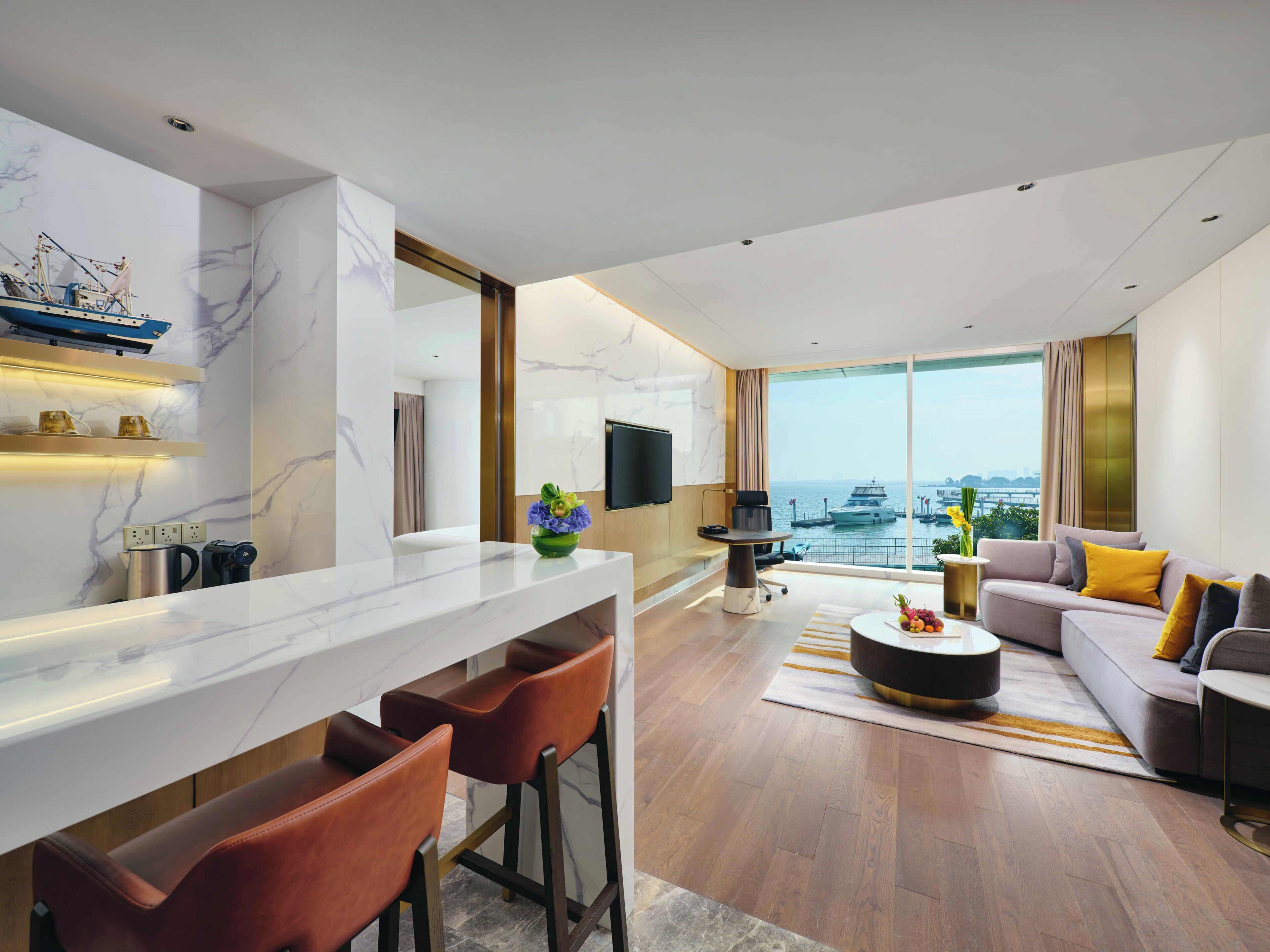 Een moderne hotelkamer met een keukenhoek, een zithoek met een bank en stoelen, een groot raam met uitzicht over de stad, en een wandgemonteerde flatscreen televisie.