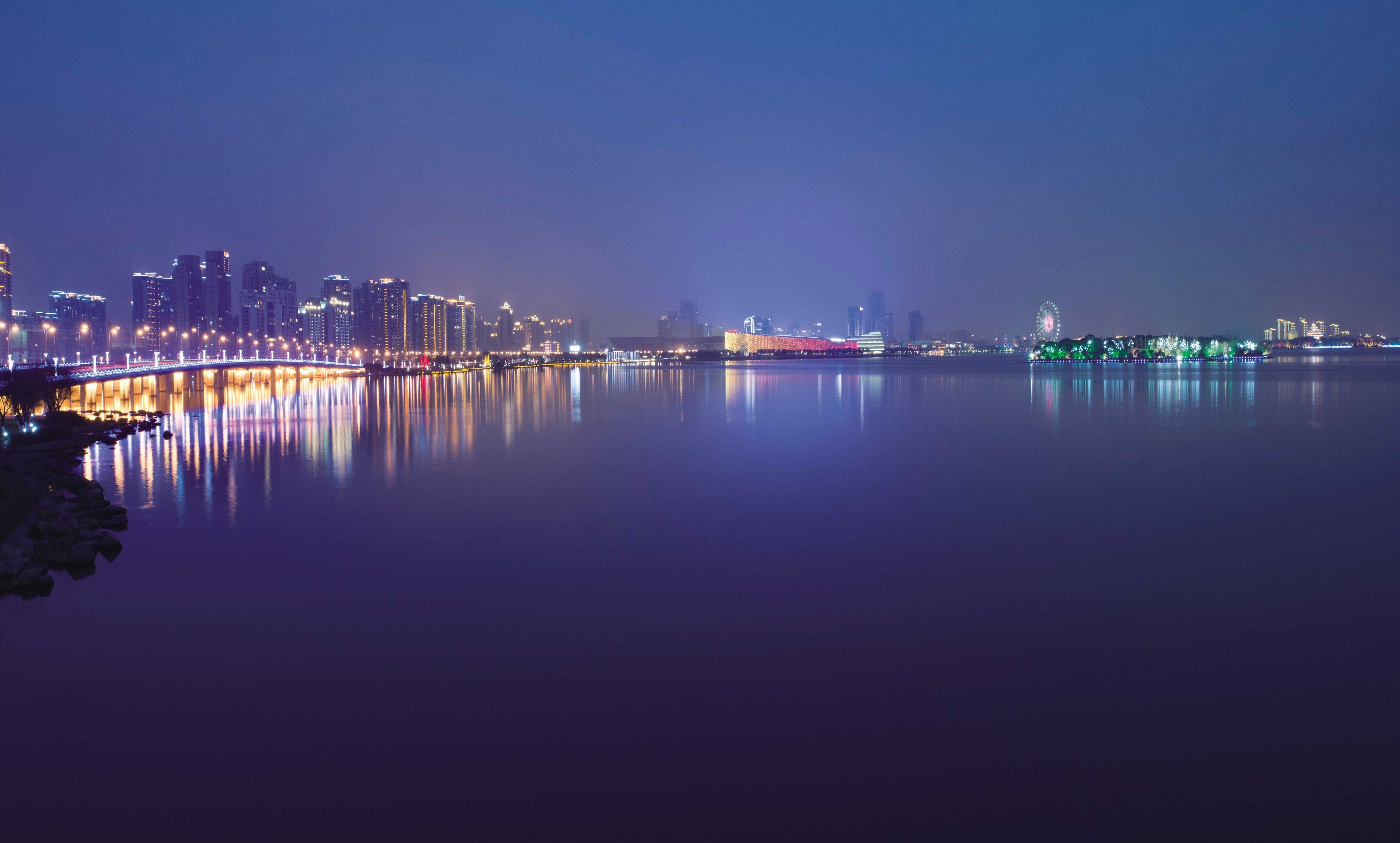 A nighttime cityscape with a waterfront, illuminated buildings, and a bridge reflecting on the water.