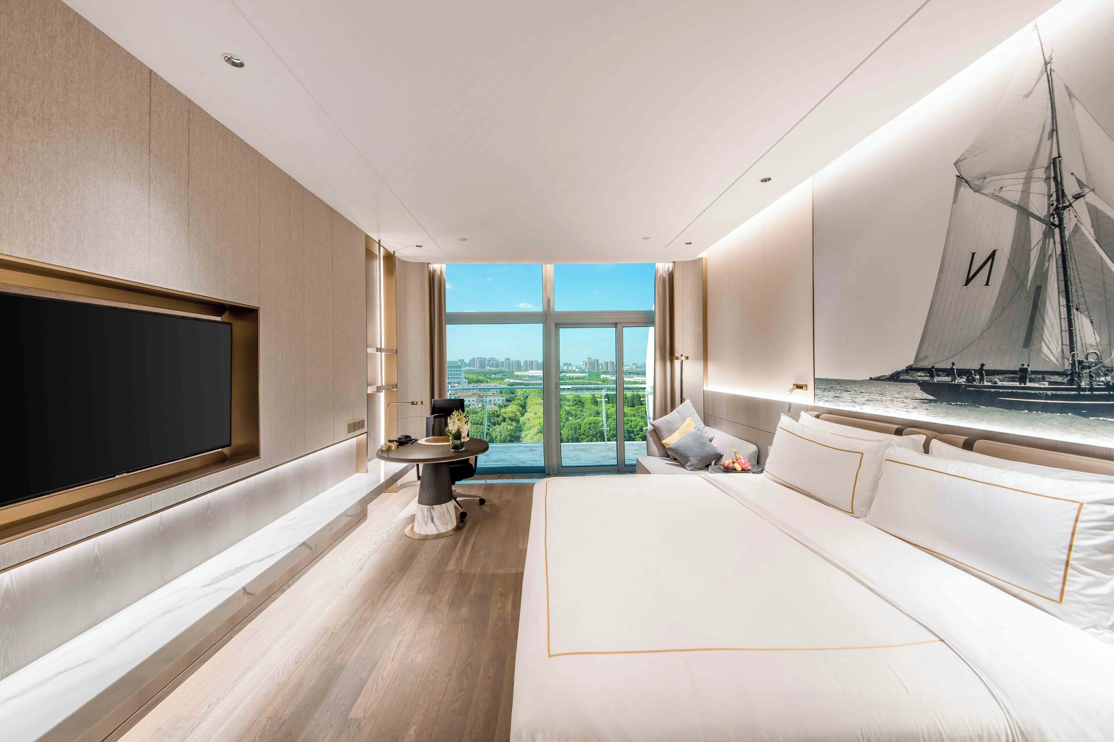 Una habitación de hotel moderna con una cama grande, un televisor de pantalla plana, una mesa de comedor y un ventanal con vistas a la ciudad.