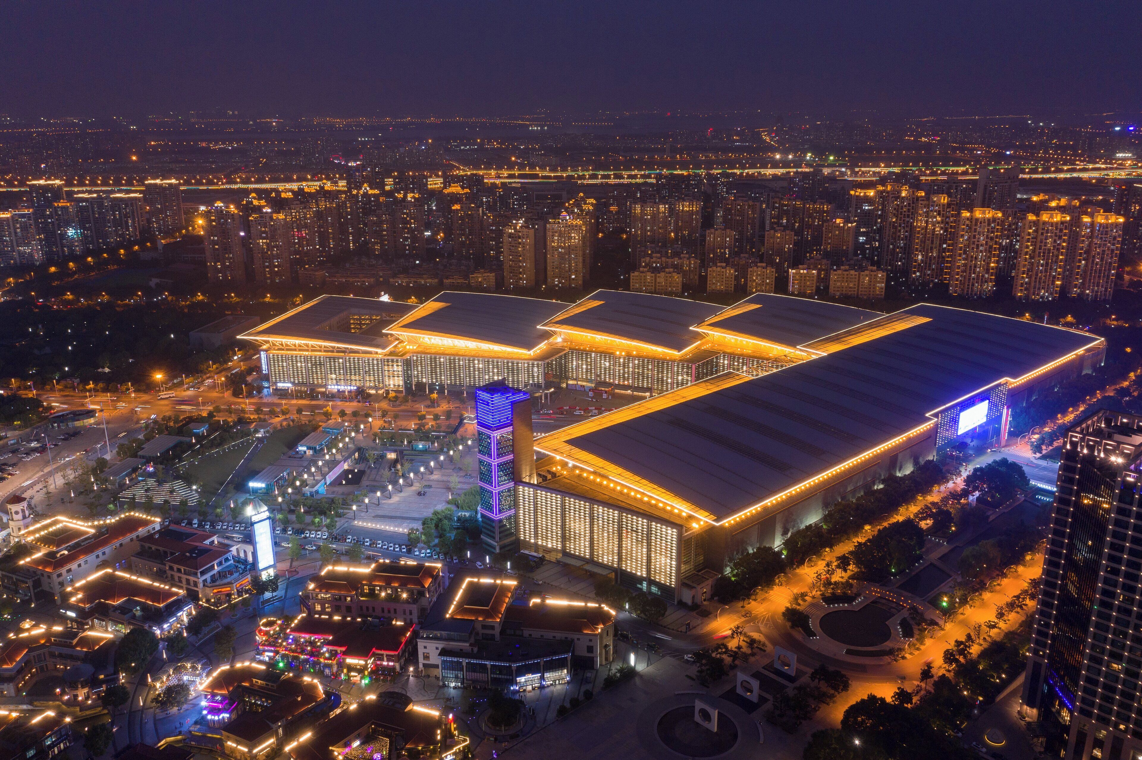 A large, multi-story building with a flat roof, surrounded by a well-lit urban area at night.