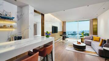 A modern hotel room with a marble kitchen island, a seating area with a sofa and chairs, a large window with a city view, and a flat-screen TV mounted on the wall.