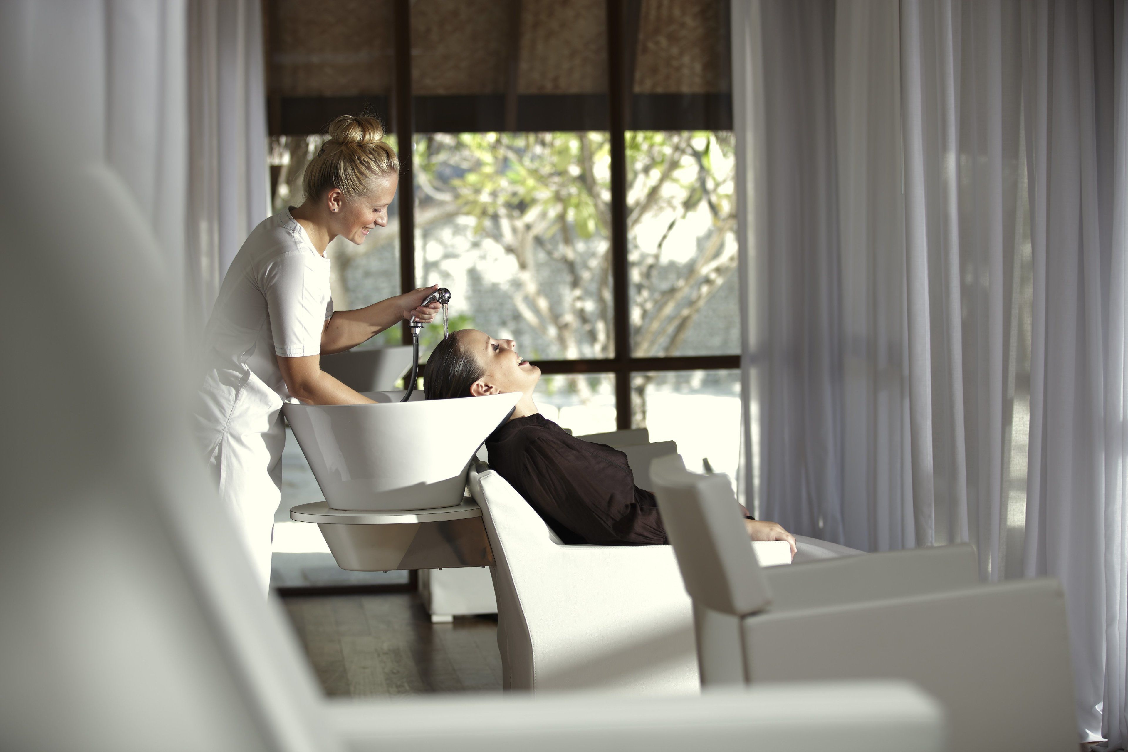 A modern room with a large window, a white chair, and a person attending to another seated individual.