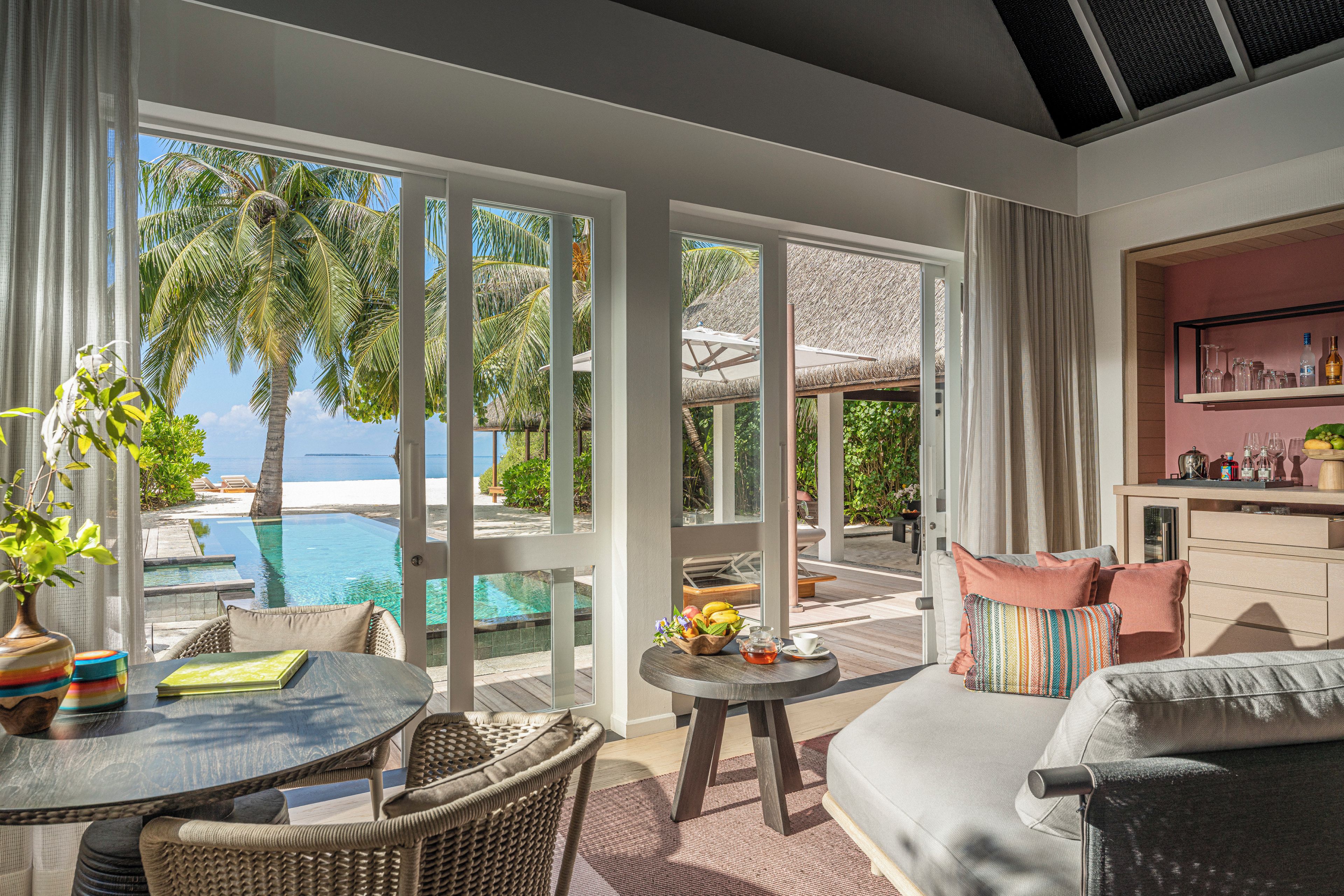 Une chambre avec vue sur une piscine et des palmiers, un coin repas avec une table et des chaises, et un espace détente avec un canapé et des coussins.